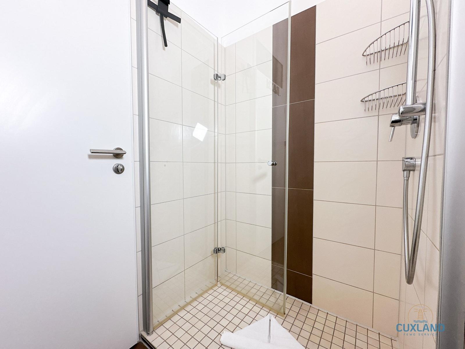 Dusche mit Glaswand, Duschbrause und Regal an der Wand.