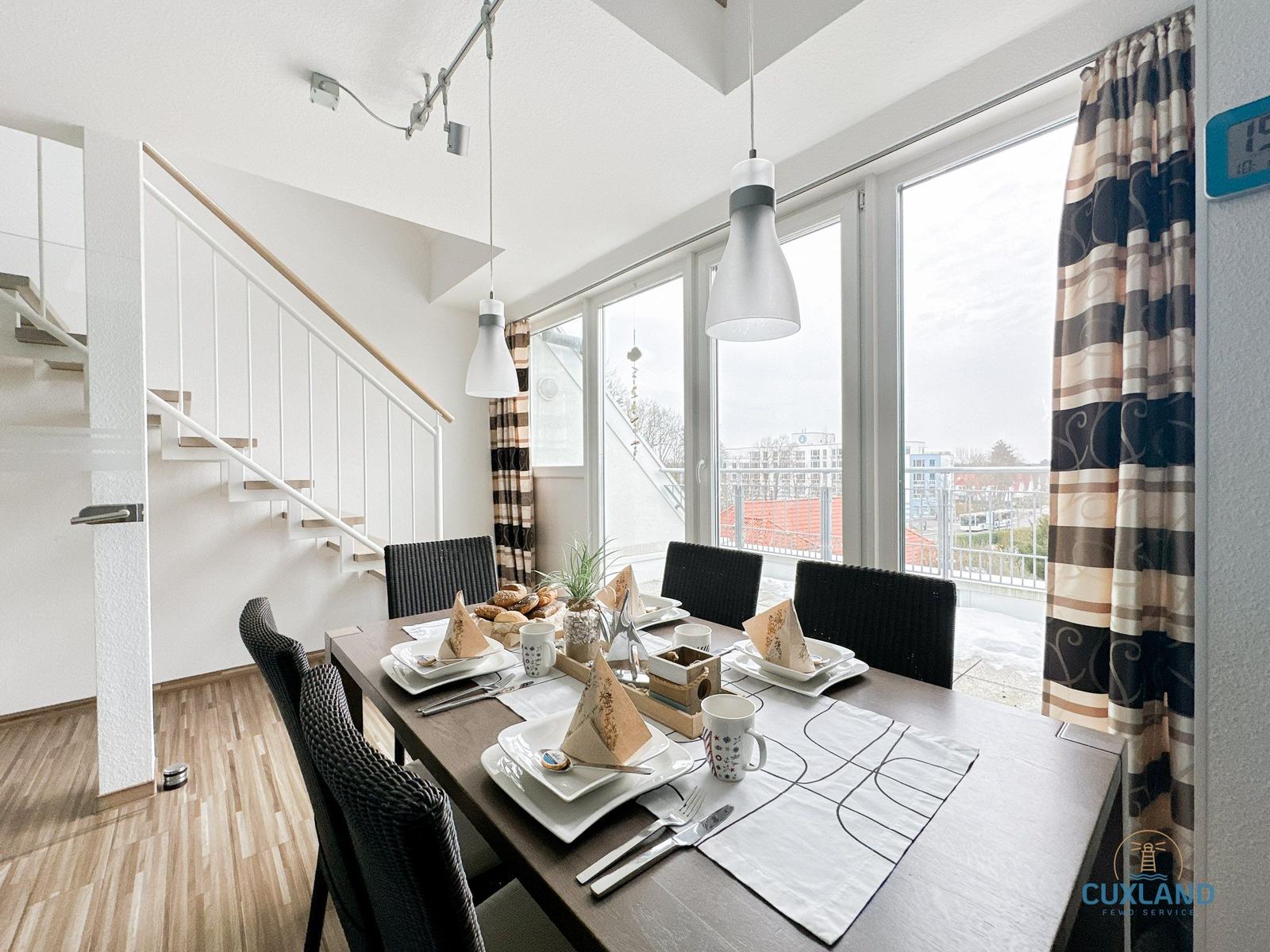 Esstisch mit sechs Stühlen und Blick auf Balkon. Helle Küche mit Treppe und Fenstern.