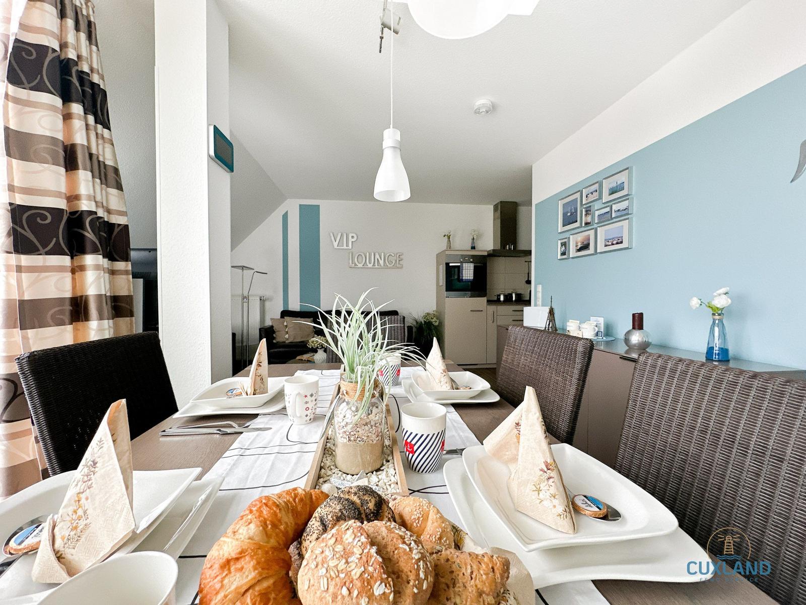 Geschmückter Esstisch mit Brot und Tassen. Im Hintergrund Küche und Wohnbereich mit Wanddekoration.