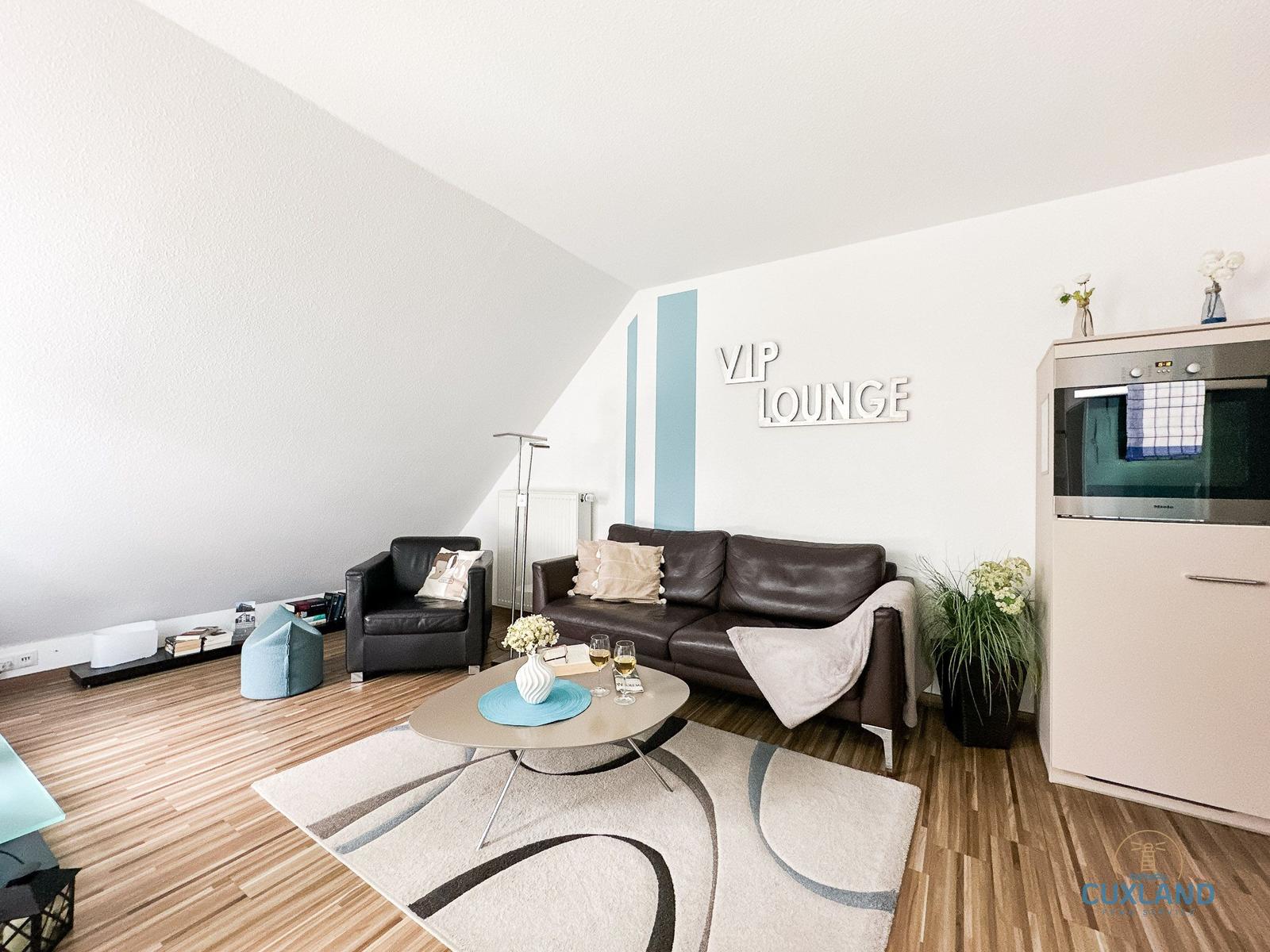 Wohnzimmer mit Sofa, Sessel und Küchenzeile. Wand mit VIP LOUNGE Beschriftung.