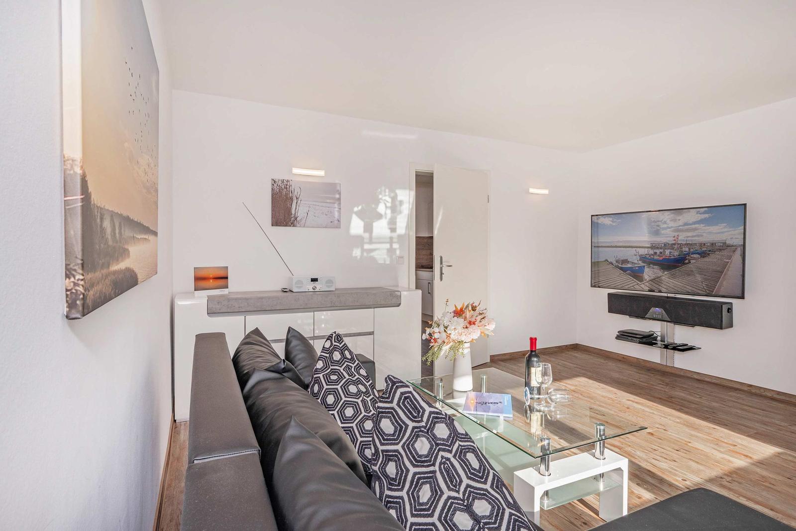 Wohnzimmer mit Sofa, Glas-Tisch und Wand-TV. Holzboden und Wandbilder.