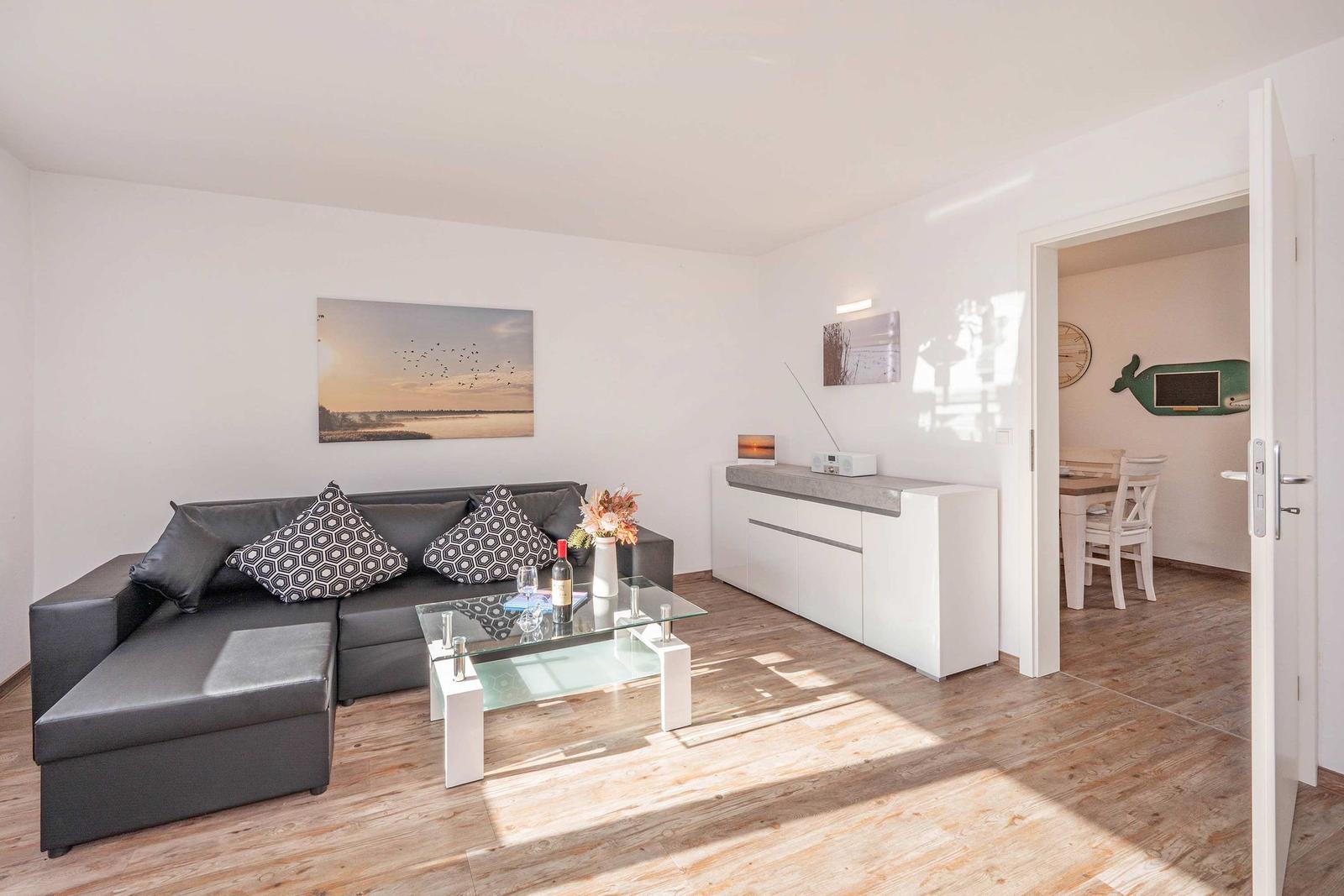 Wohnzimmer mit grauem Sofa, Glas-Tisch und Küchenbereich im Hintergrund.