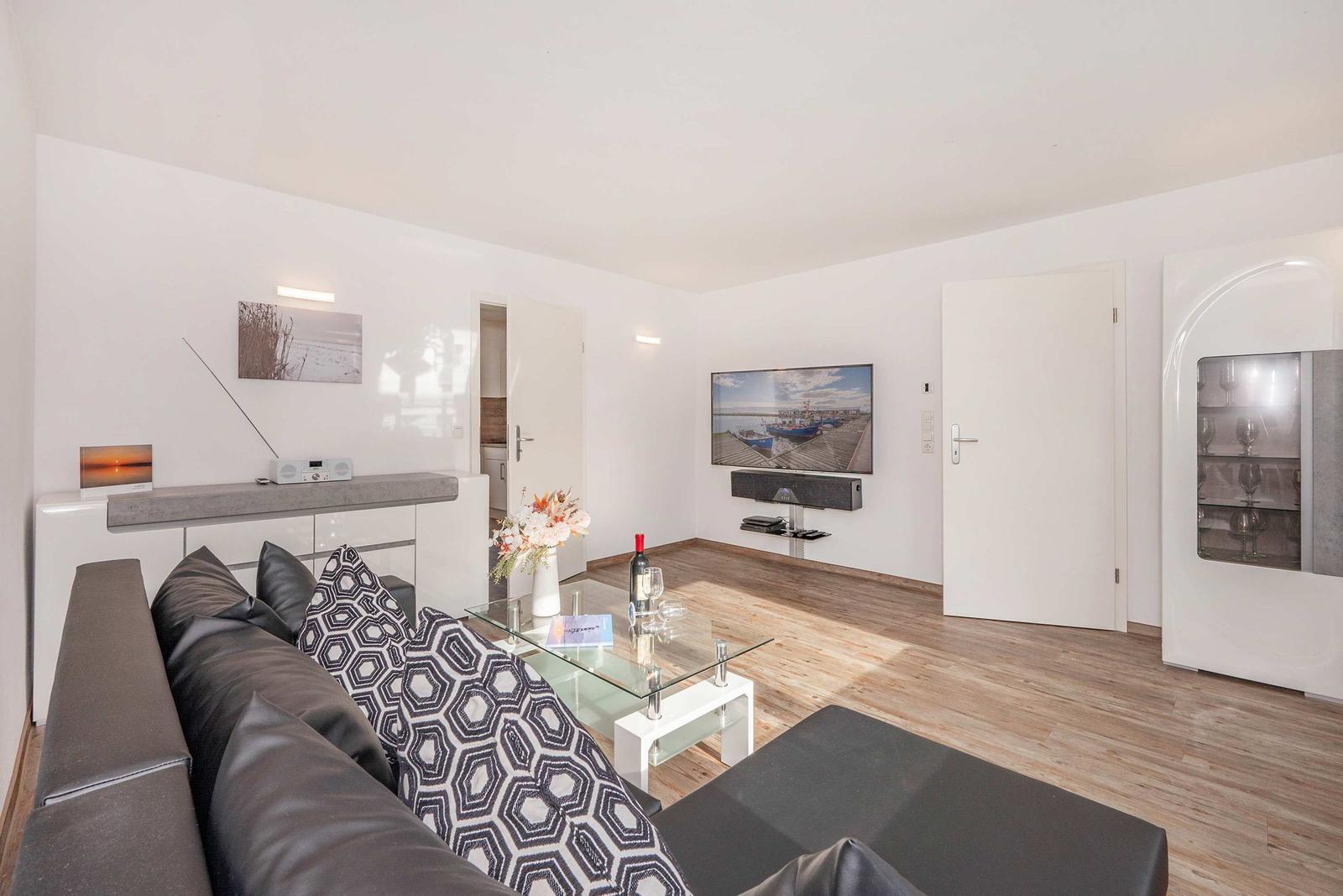 Wohnzimmer mit Sofa, Glas-Tisch und Wandfernseher. Holzboden und weiße Wände.