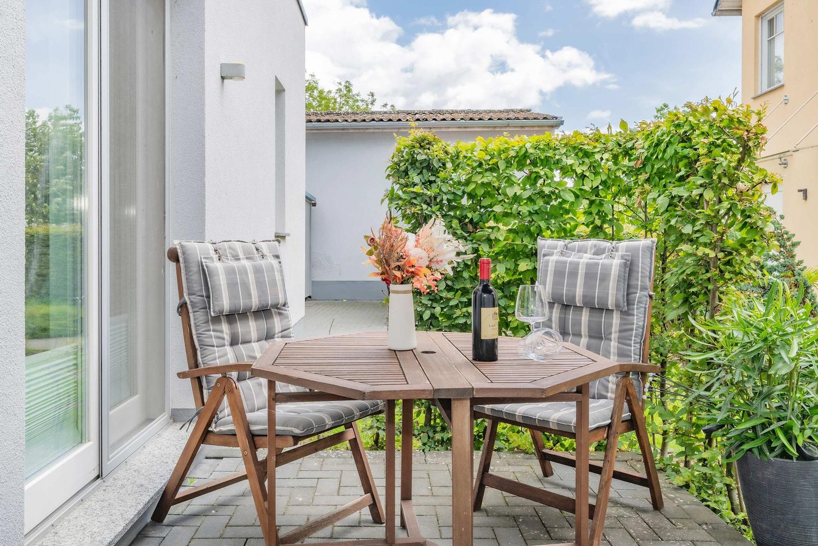 Terrasse mit Holztisch, zwei Stühlen und einer Weinflasche.