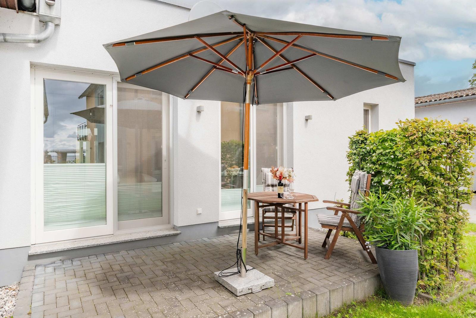 Terrasse mit Sonnenschirm, Tisch und Stühlen. Hintergrund: Haus mit Fenstern und Beete.