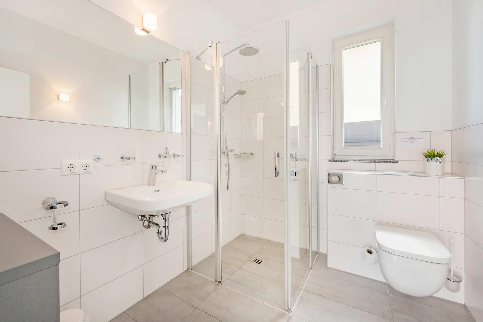 Badezimmer mit Dusche, Waschbecken und WC. Weiße Fliesen und Fenster.