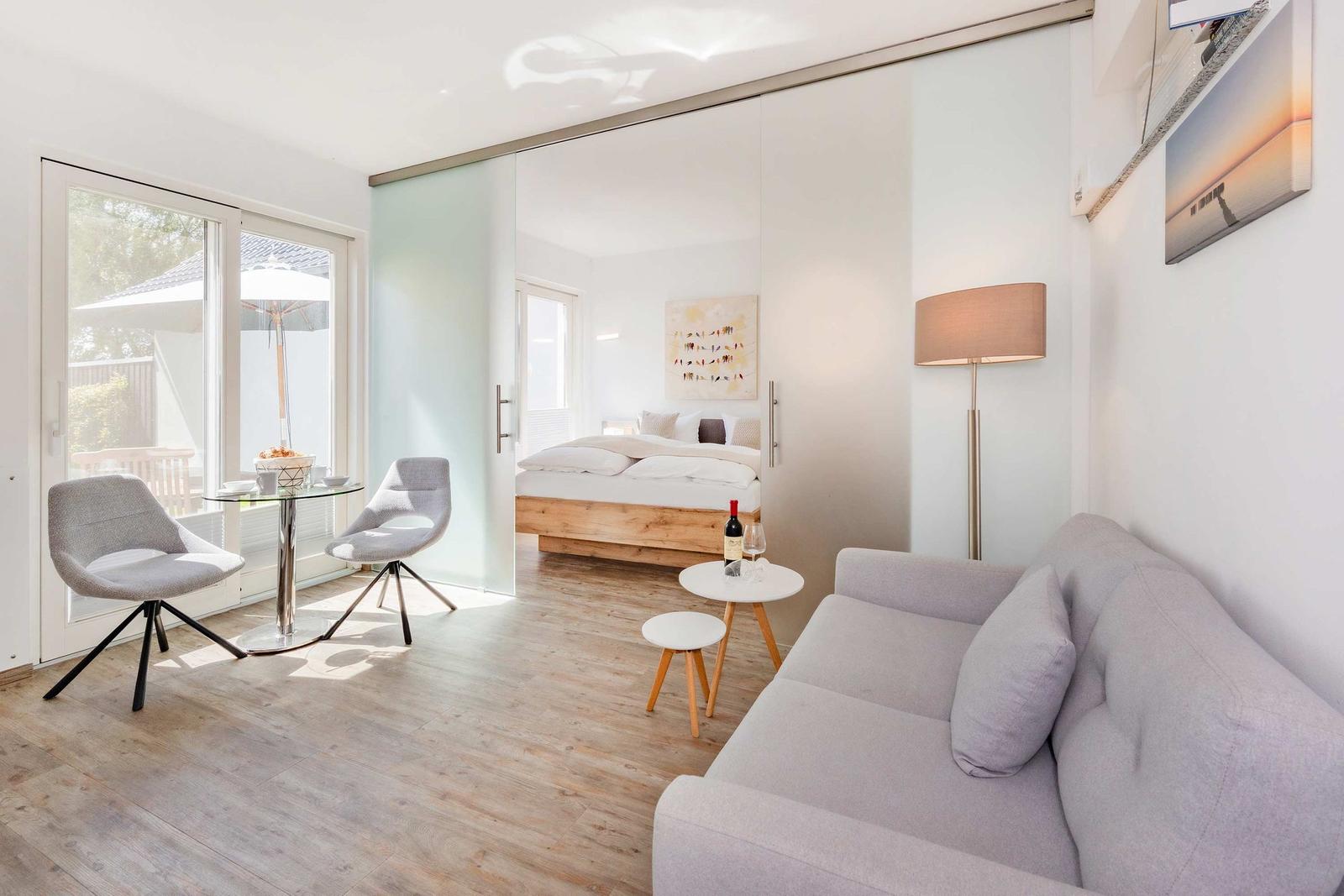 Wohnbereich mit Sofa, Tisch und Stühlen. Schlafzimmer mit Bett hinter Glaswand sichtbar.