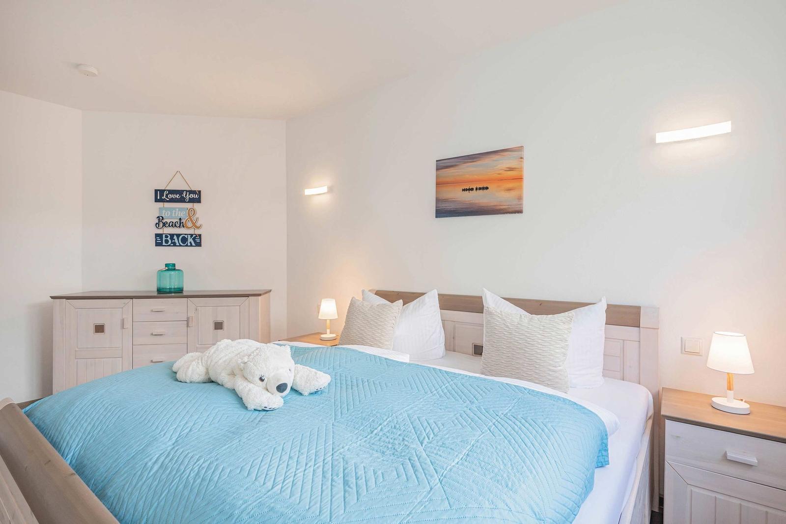 Bedroom with bed, nightstands, and wall art. A stuffed bear lies on the bed.