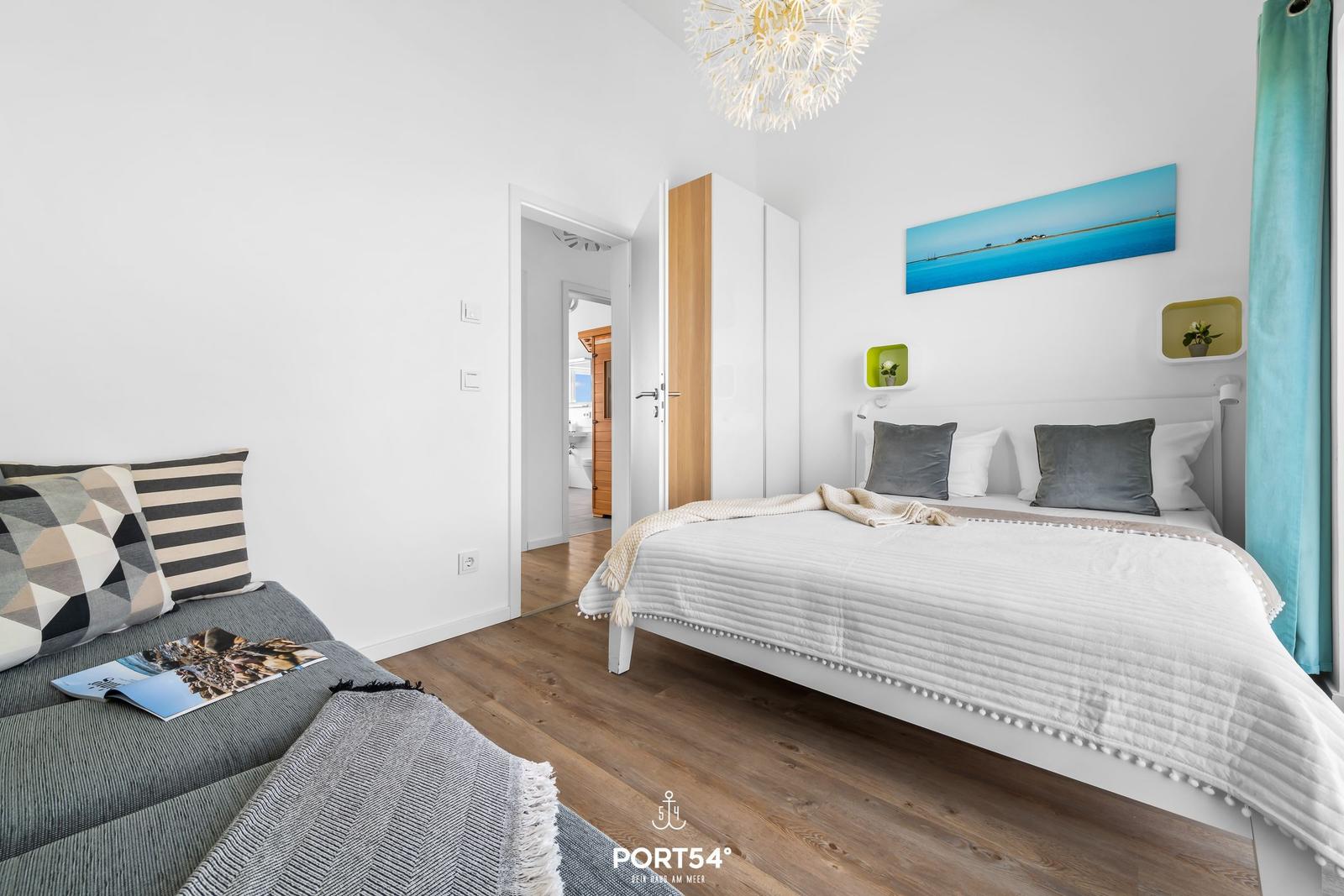 Schlafzimmer mit Bett, Sofa und Holzboden. Wandbild und Deckenleuchte sichtbar.