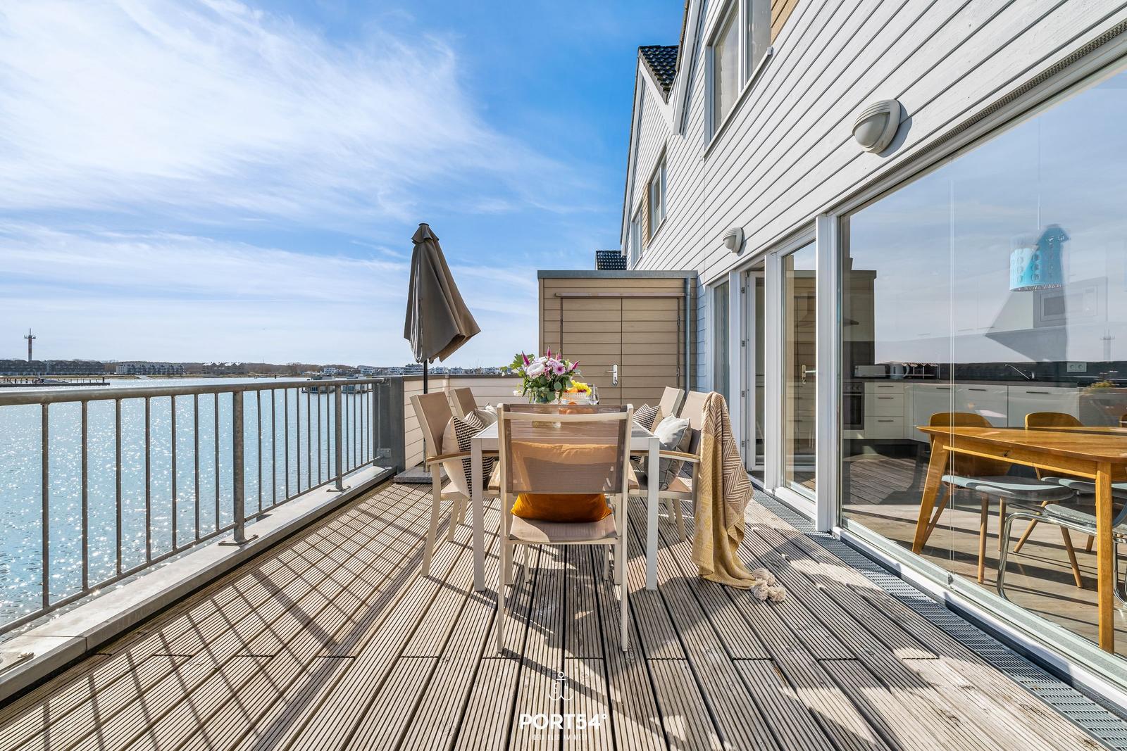 Terrasse mit Tisch, Stühlen und Blick auf Wasser. Glasfront führt ins Innere.