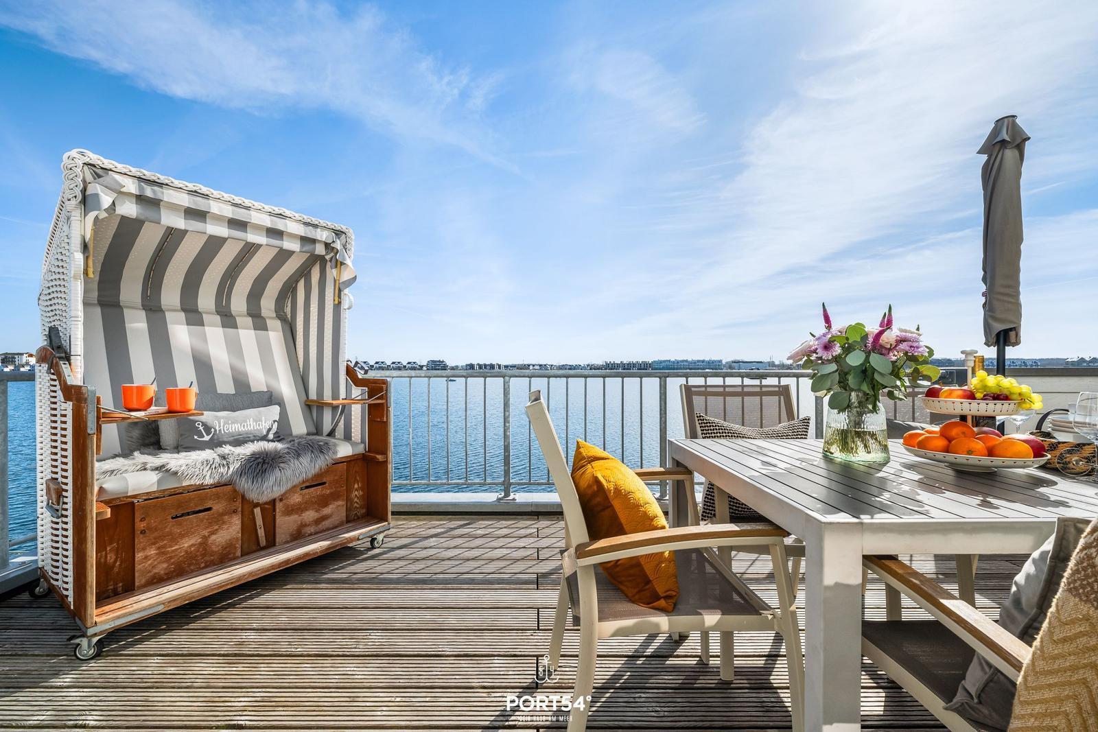 Terrasse mit Strandkorb, Tisch und Stühlen, Blick auf Wasser und Hafen.