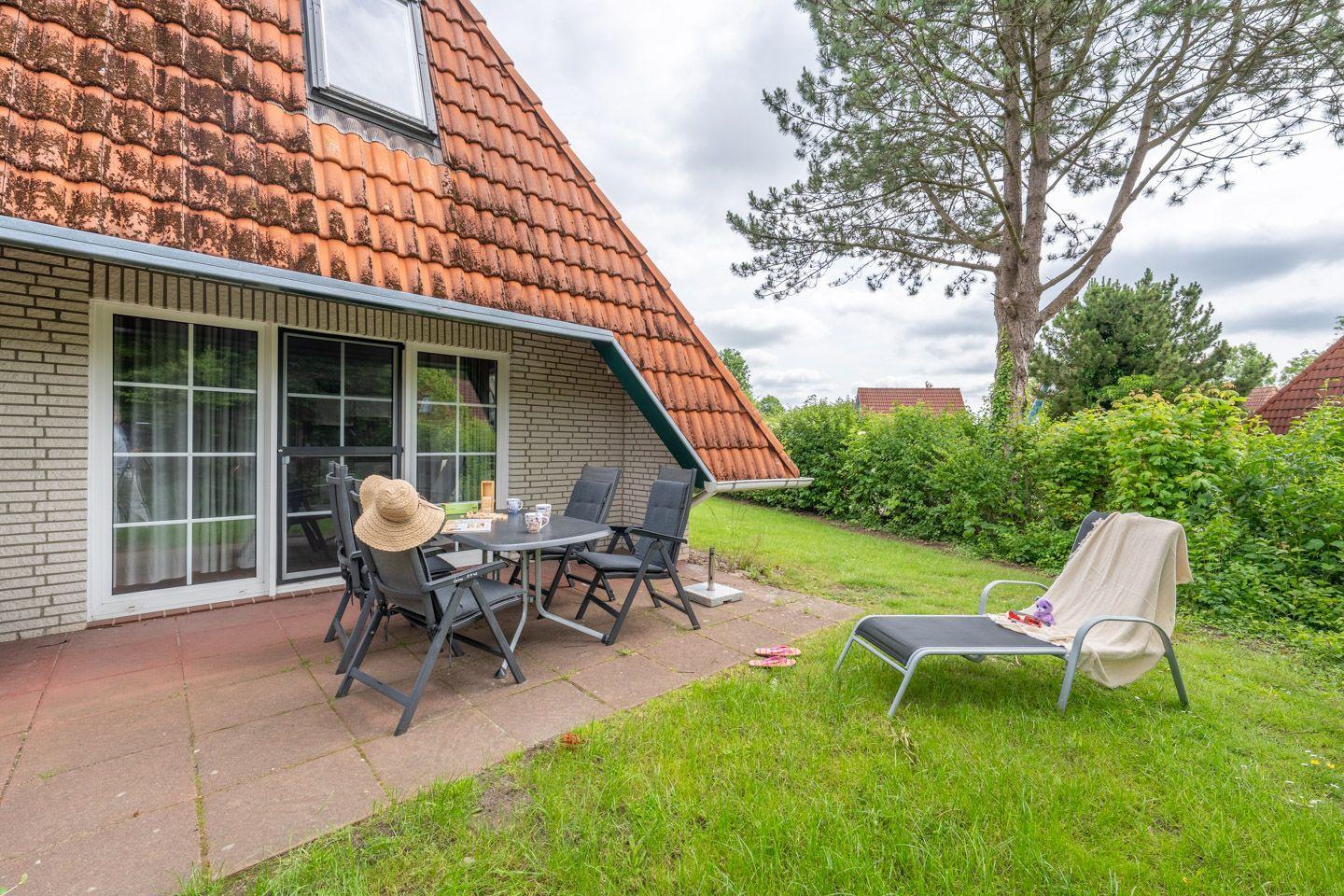 Terrasse mit Tisch, Stühlen und Liege im Garten. Hintergrund: Haus mit rotem Dach und Baum.