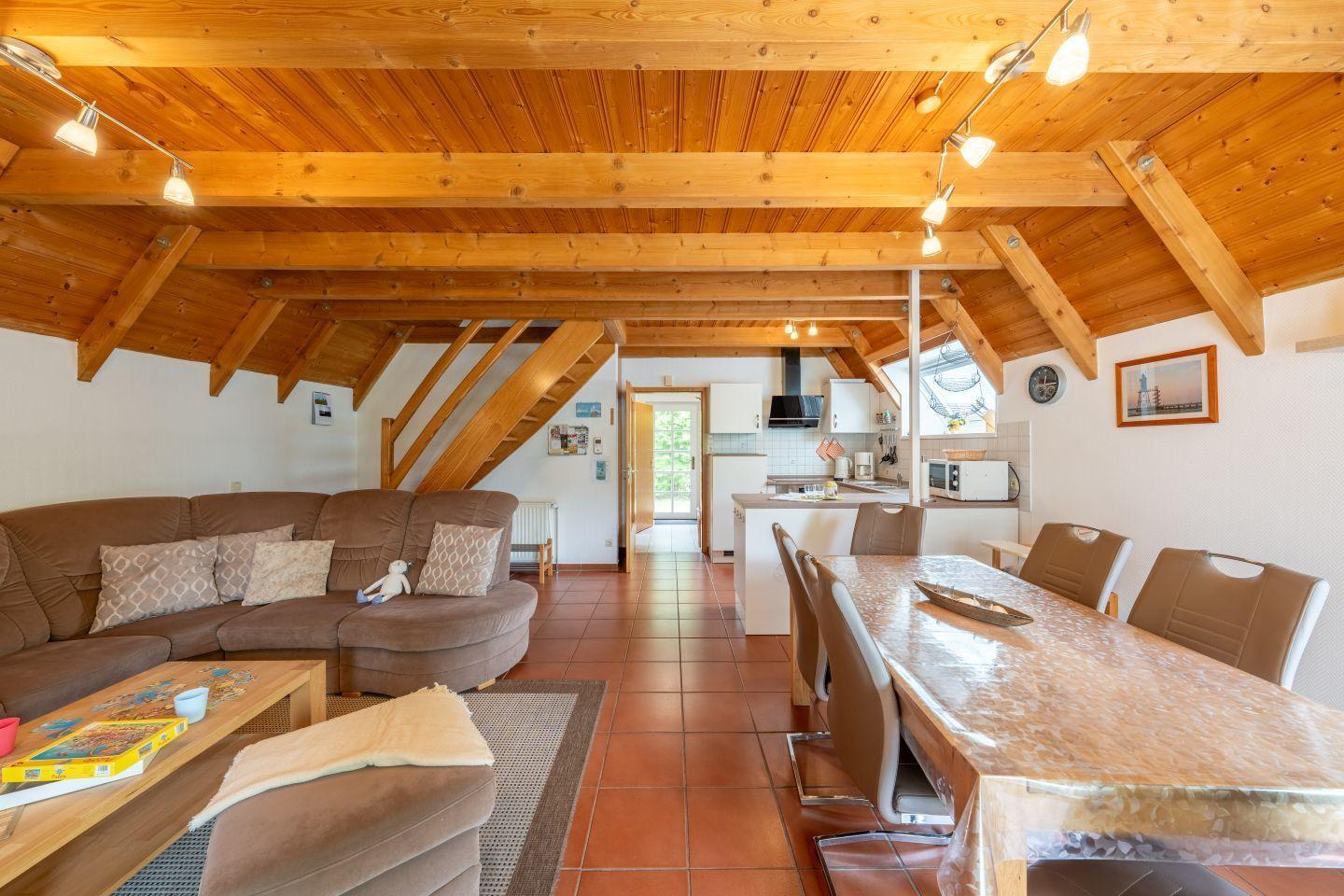 Living and dining area with wooden ceiling, sofa, dining table, and kitchen in the background.