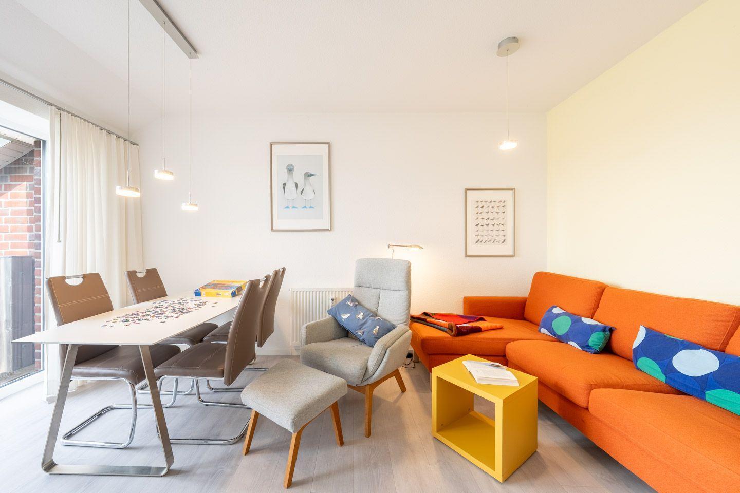 Wohn- und Essbereich mit orangefarbenem Sofa, Esstisch und Stühlen sowie Wandbildern.