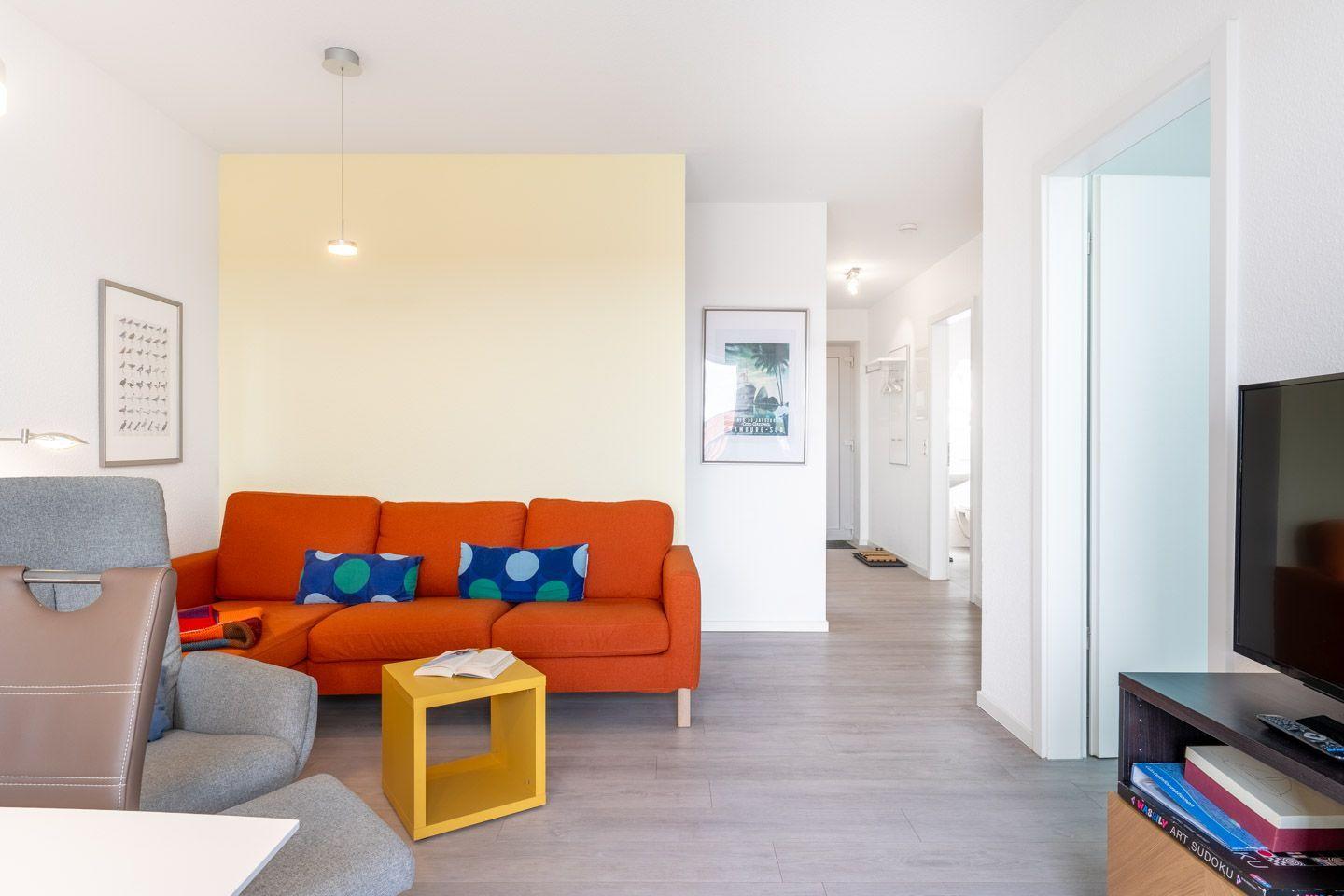 Wohnbereich mit orangefarbenem Sofa, gelbem Beistelltisch und Fernseher.