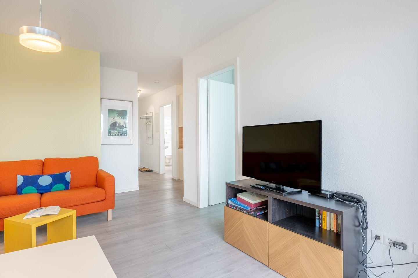 Wohnzimmer mit orangefarbenem Sofa, Fernseher und Holzmöbeln. Blick in den Flur.