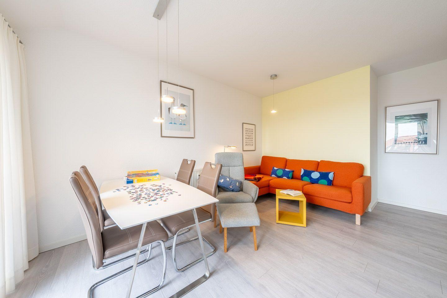 Essbereich mit Tisch und Stühlen. Wohnbereich mit orangefarbenem Sofa und gelbem Beistelltisch.