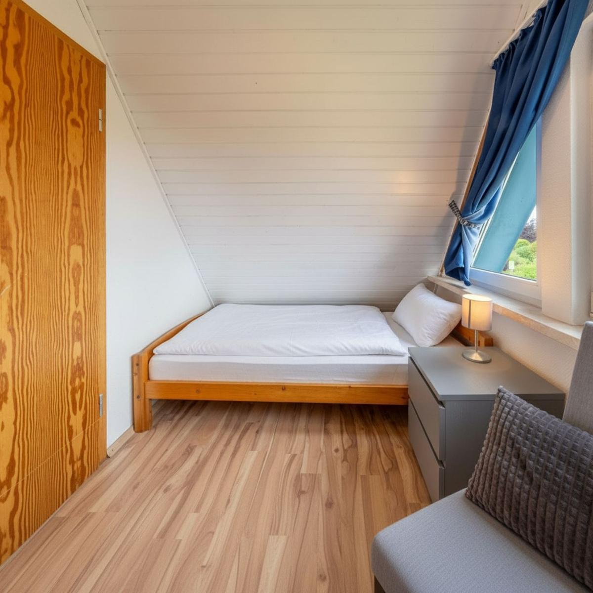 Schlafzimmer mit Holzboden, Dachschräge, Bett und Fenster mit blauen Vorhängen.