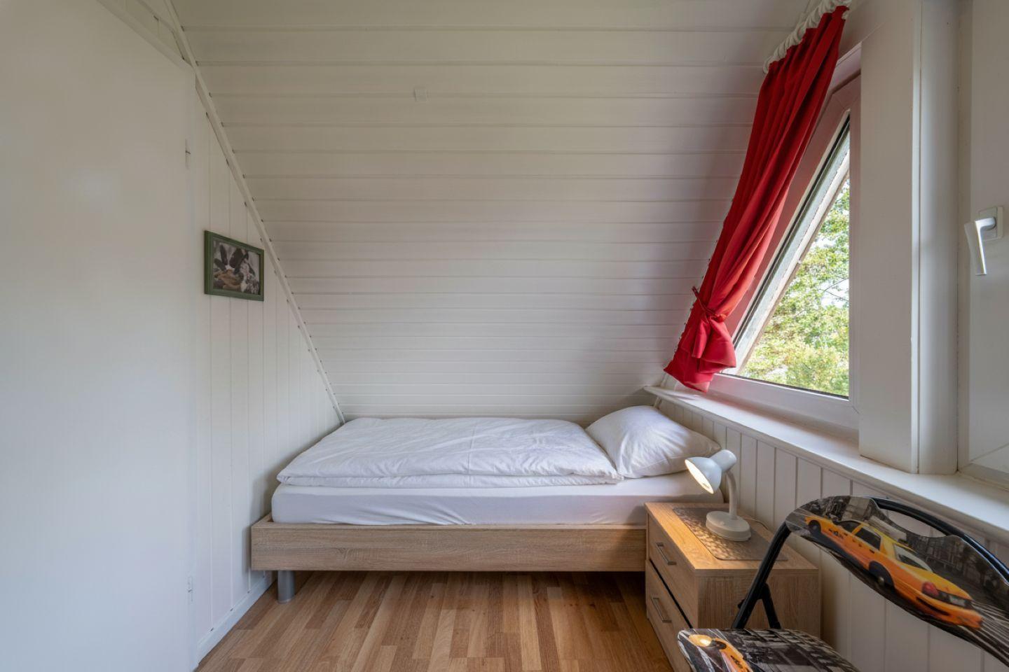 A bedroom with sloped ceiling, bed, nightstand, and window with red curtain.