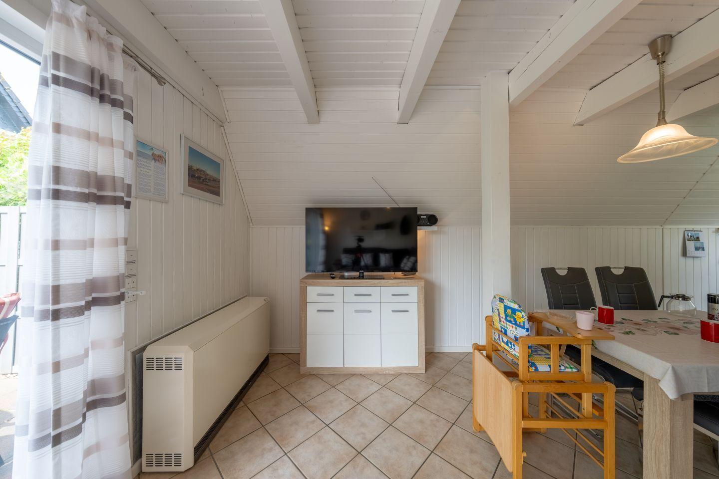 Living and dining area with TV, table and chairs, white paneled walls and wooden ceiling.