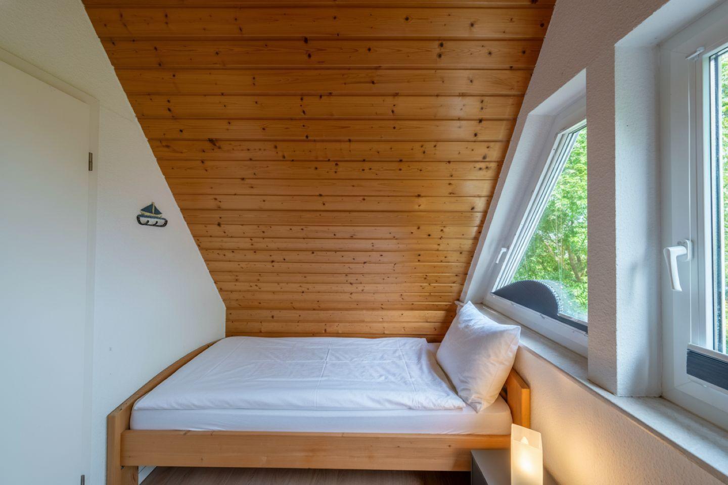 Ein Schlafzimmer mit Holzdecke, Bett und Fenster. Draußen sind Bäume sichtbar.