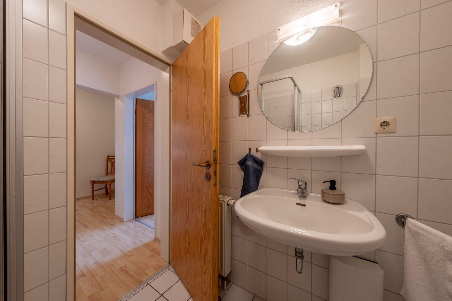 Badezimmer mit Waschbecken, Spiegel und Dusche. Tür führt in Wohnbereich.