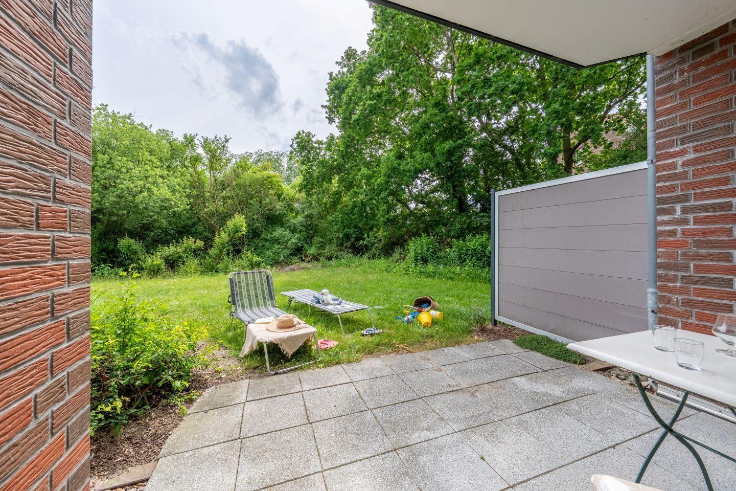 Terrace with table, chair, and garden. Background: green area and trees.