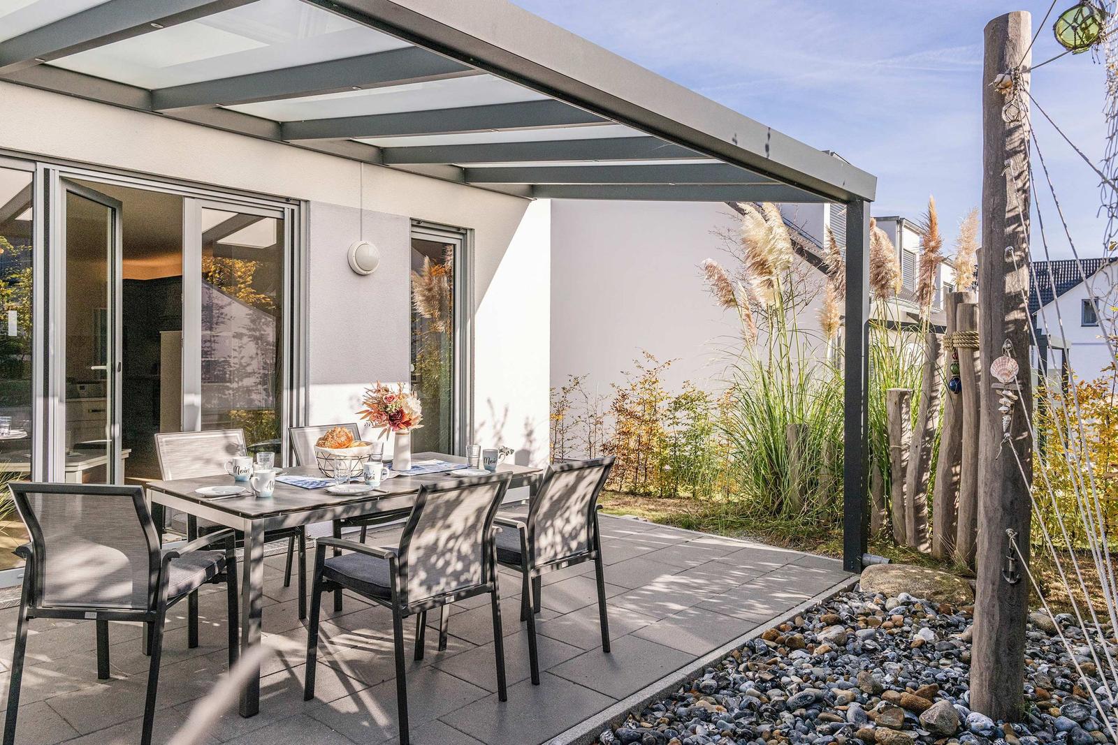 Terrasse mit Tisch, Stühlen und Markise. Hintergrund: Grünflächen und Steinbeete.