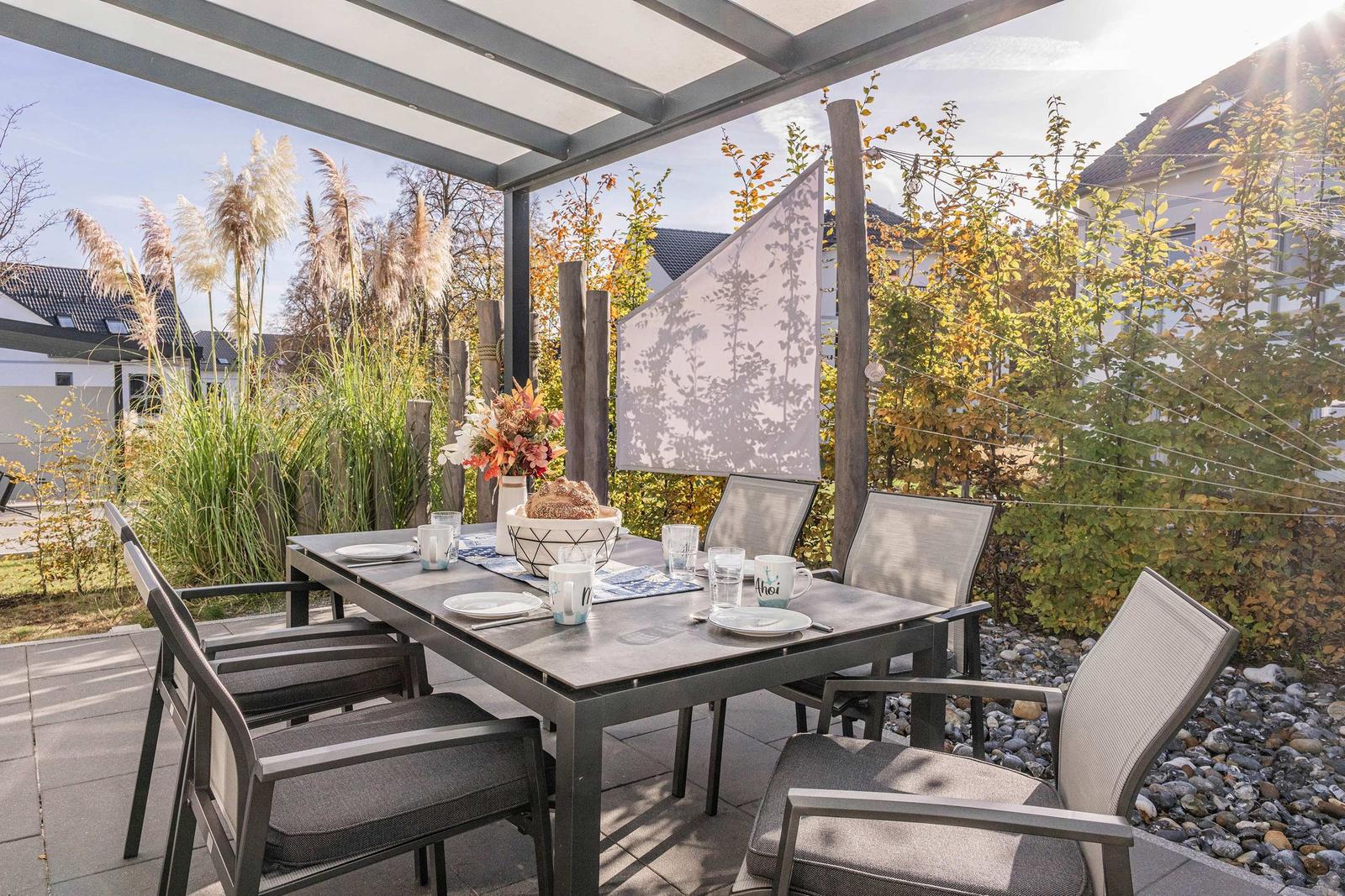 Terrasse mit gedecktem Tisch und Stühlen unter Pavillon. Umgeben von Pflanzen und Steinchen.