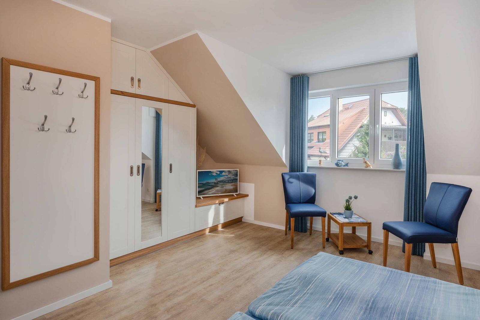 Schlafzimmer mit Dachschräge, Kleiderkabine, TV und zwei blauen Stühlen vor Fenster.