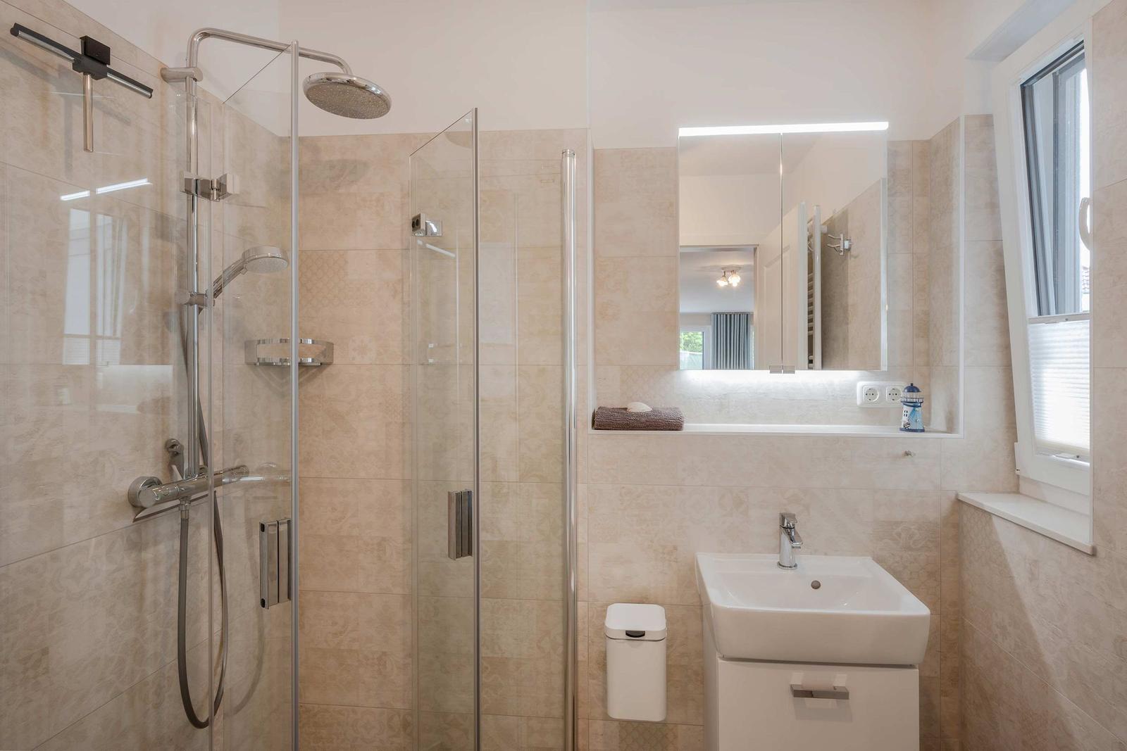 Bad mit Dusche, Waschbecken und Spiegel. Fenster sorgt für Tageslicht.