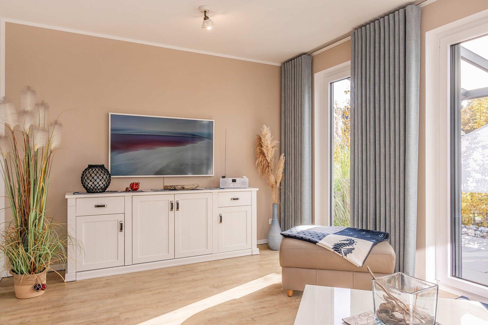 Wohnzimmer mit TV, weißen Schränken, grauen Vorhängen und Blick auf den Garten.
