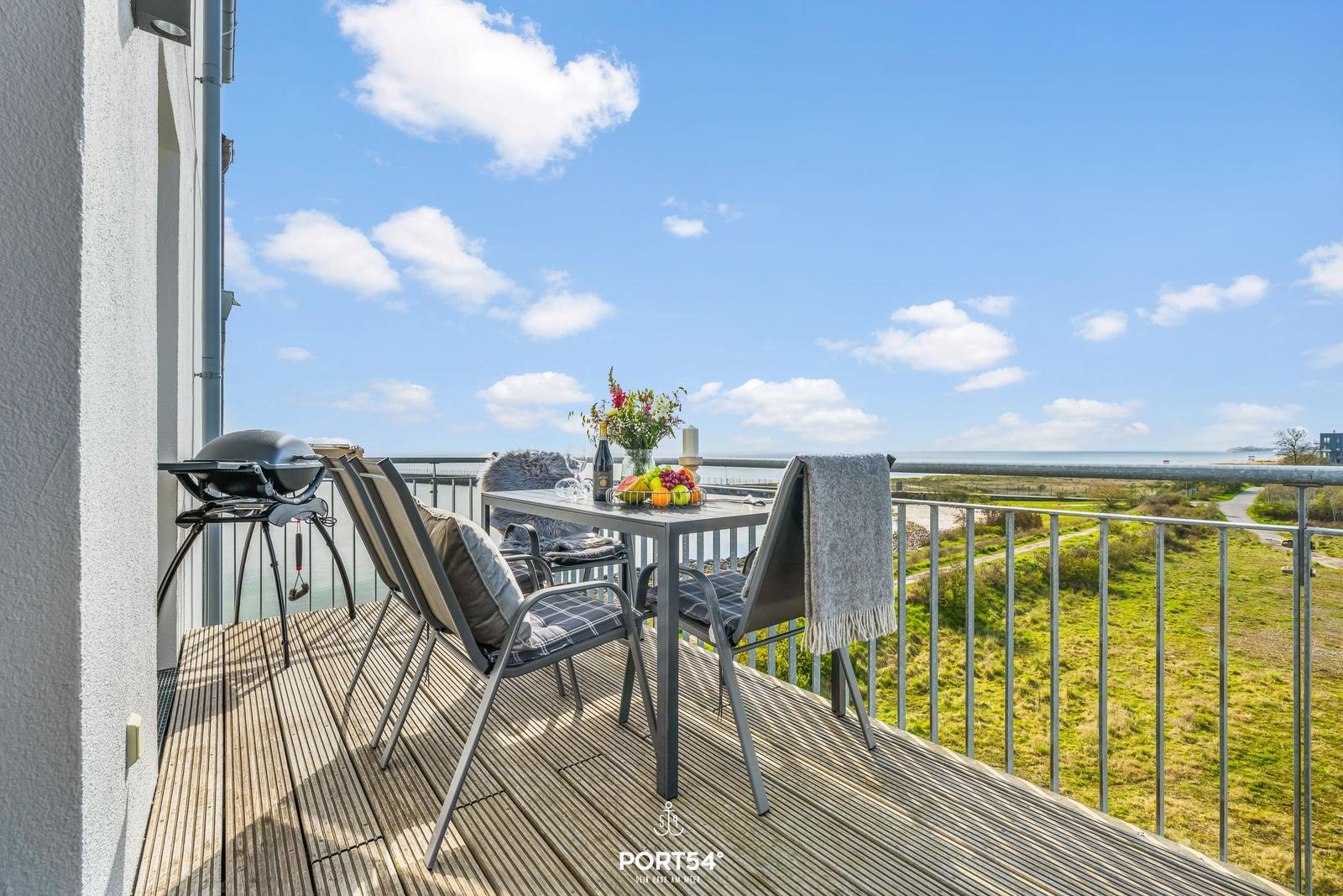 Terrasse mit Grill, Tisch und Stühlen. Blick auf Meer und Grünfläche.