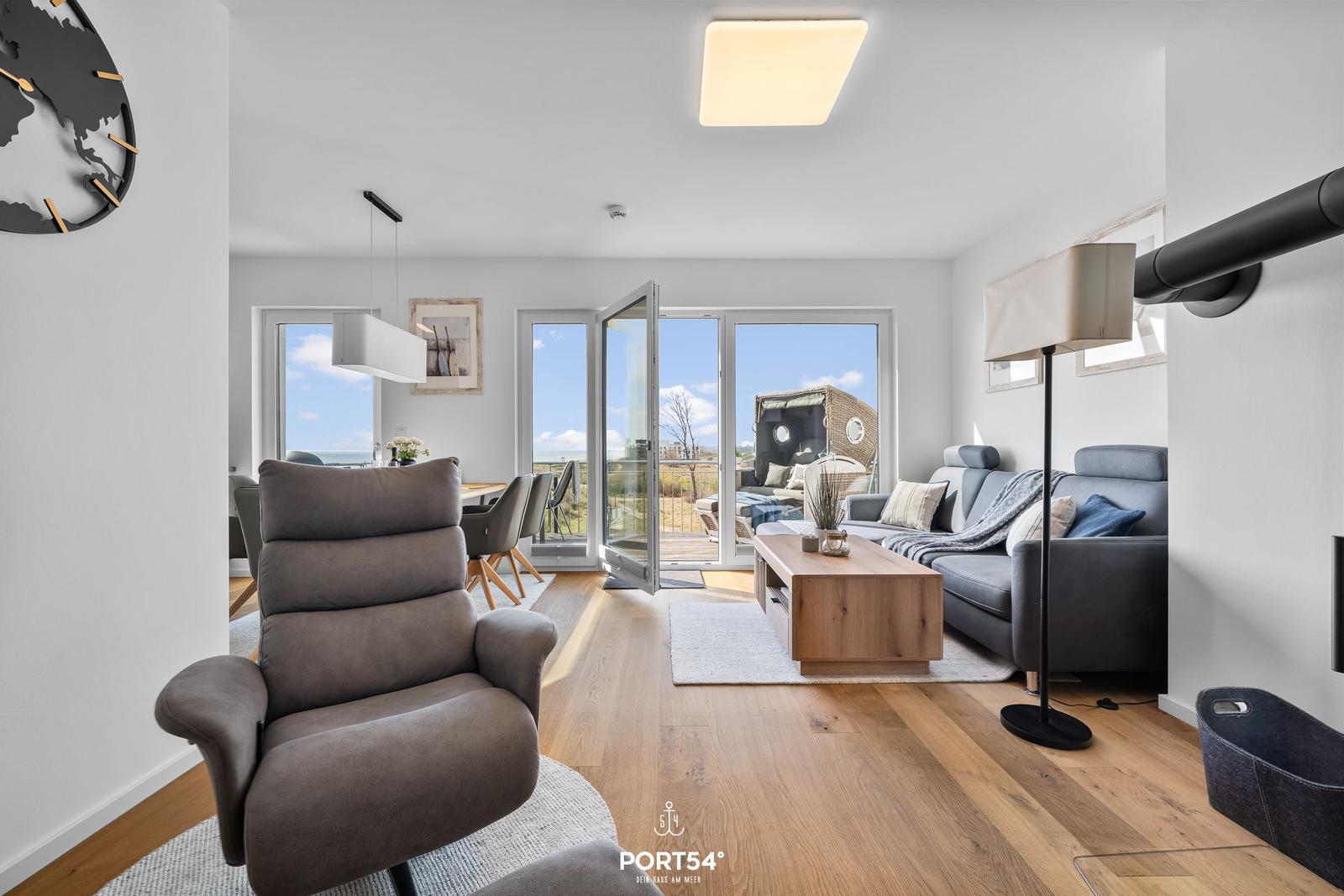 Wohnzimmer mit Sitzgruppe, Holzboden und Blick auf Balkon. Tischlampe und Wanduhr sichtbar.