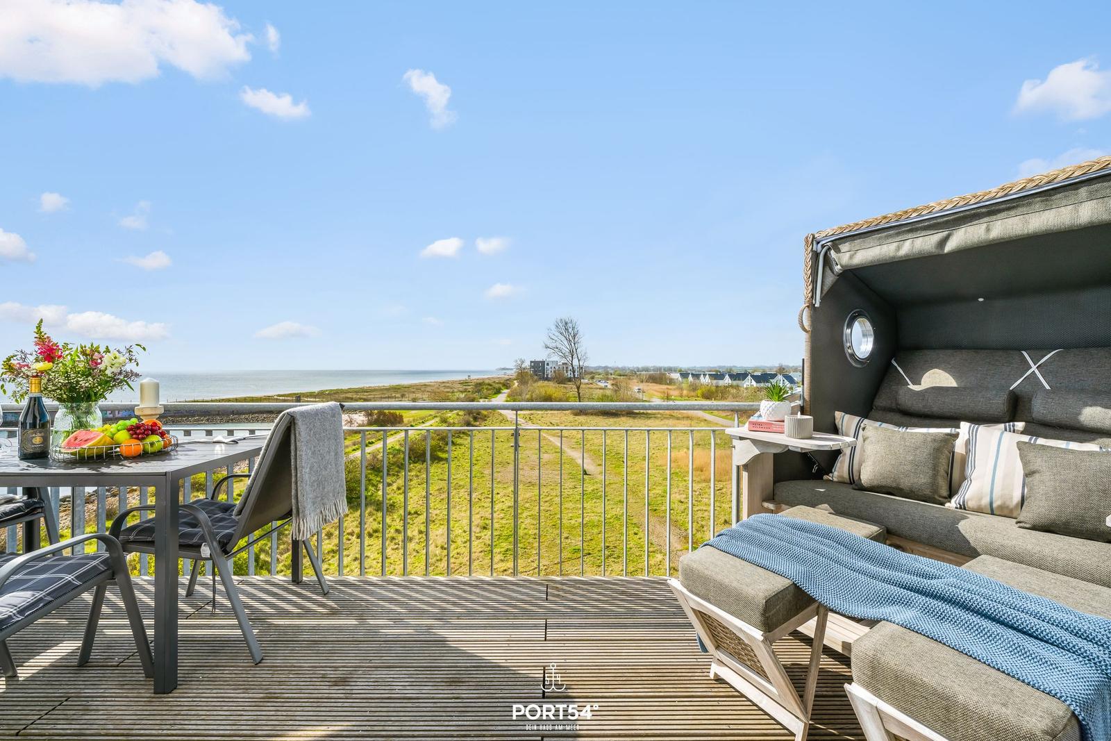 Terrasse mit Liege, Tisch und Meerblick. Himmel blau, Grasland und Wasser sichtbar.