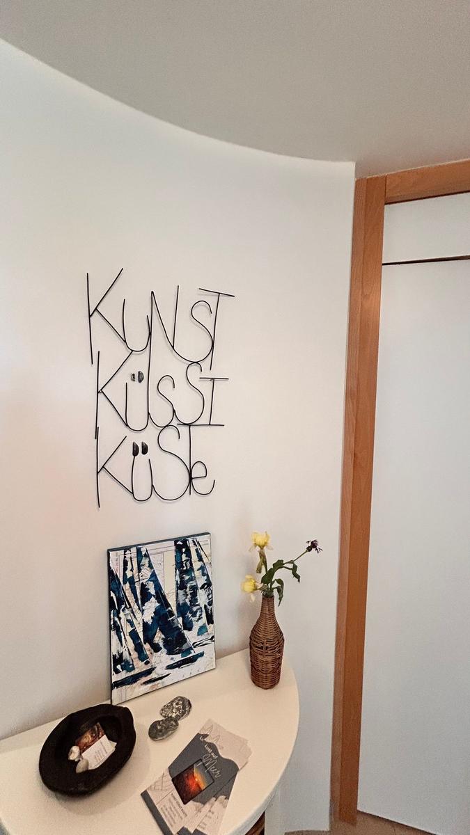 Wanddekoration mit Schriftzug, Bild und Blumen in Vase auf runder Tischkante.