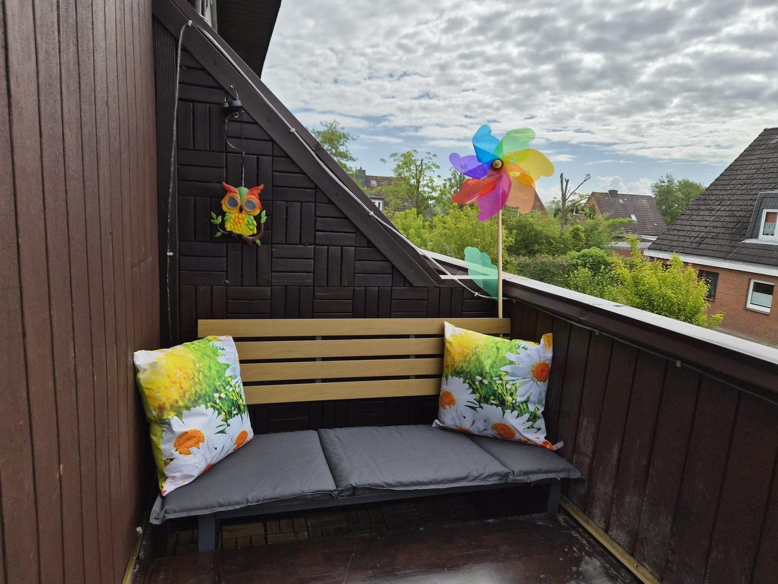 Terrasse mit Bank, Kissen und Windrad. Blick auf Grün und Häuser.