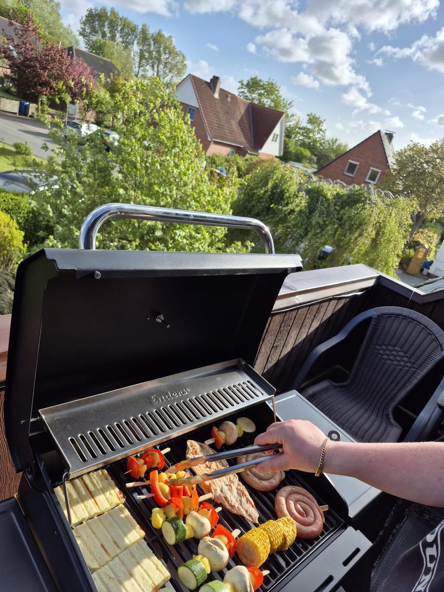Grill mit Fleisch und Gemüse auf Terrasse mit Sicht auf Grün und Häuser.