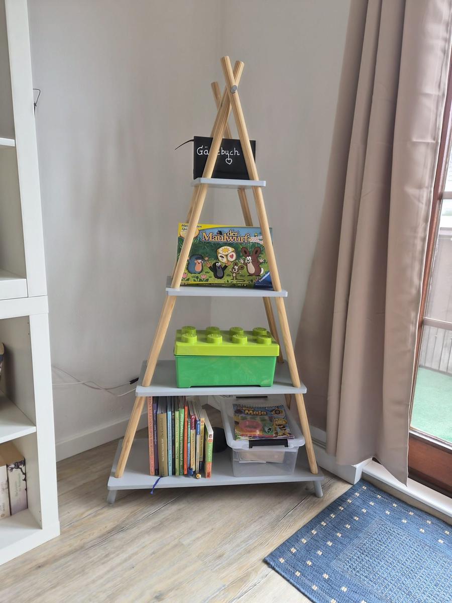 Dreieckiger Holzregal mit Büchern, Spielzeug und Spielboxen im Raum.