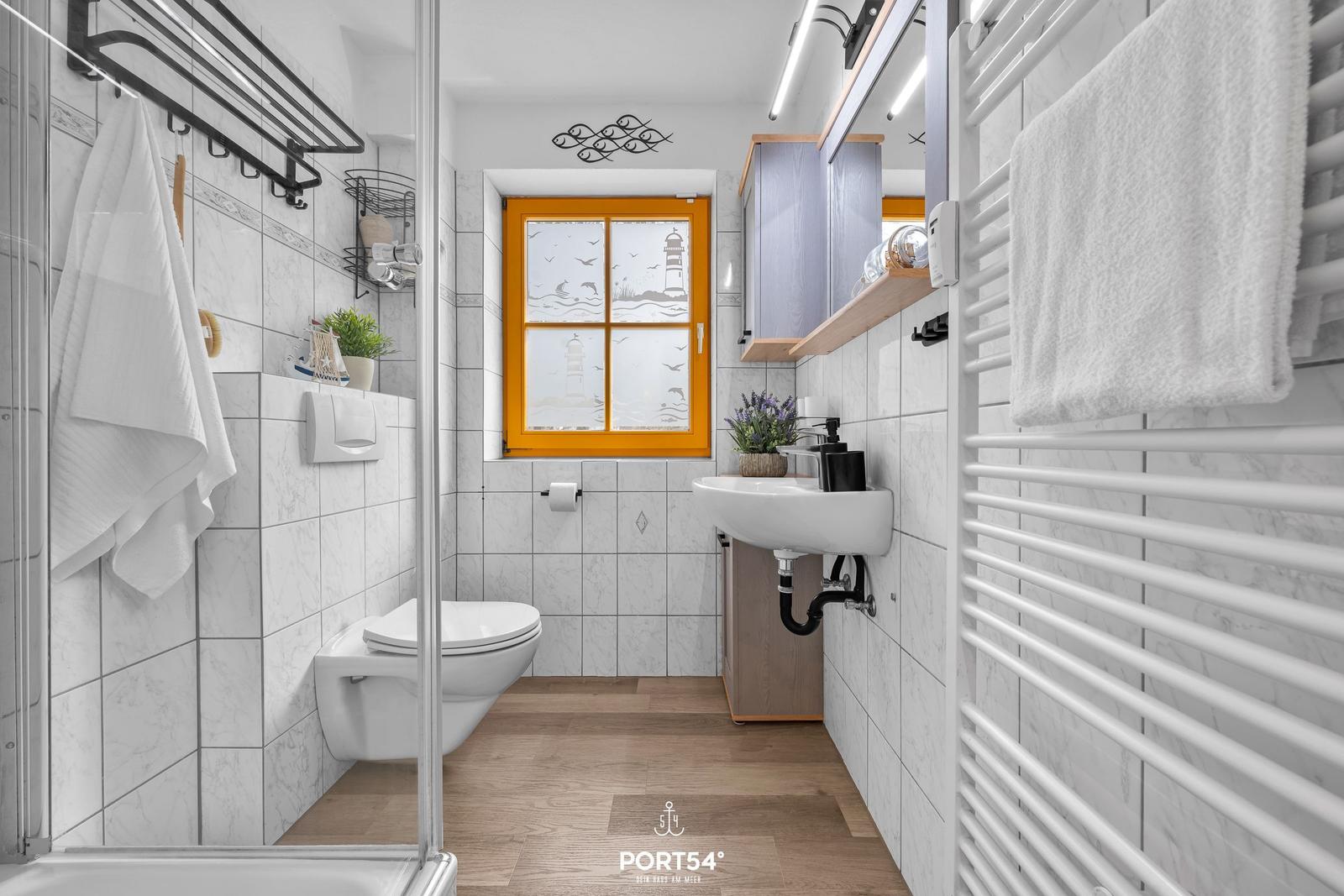 Badezimmer mit Toilette, Waschbecken und Dusche. Fenster mit Leuchtturm-Motiv.