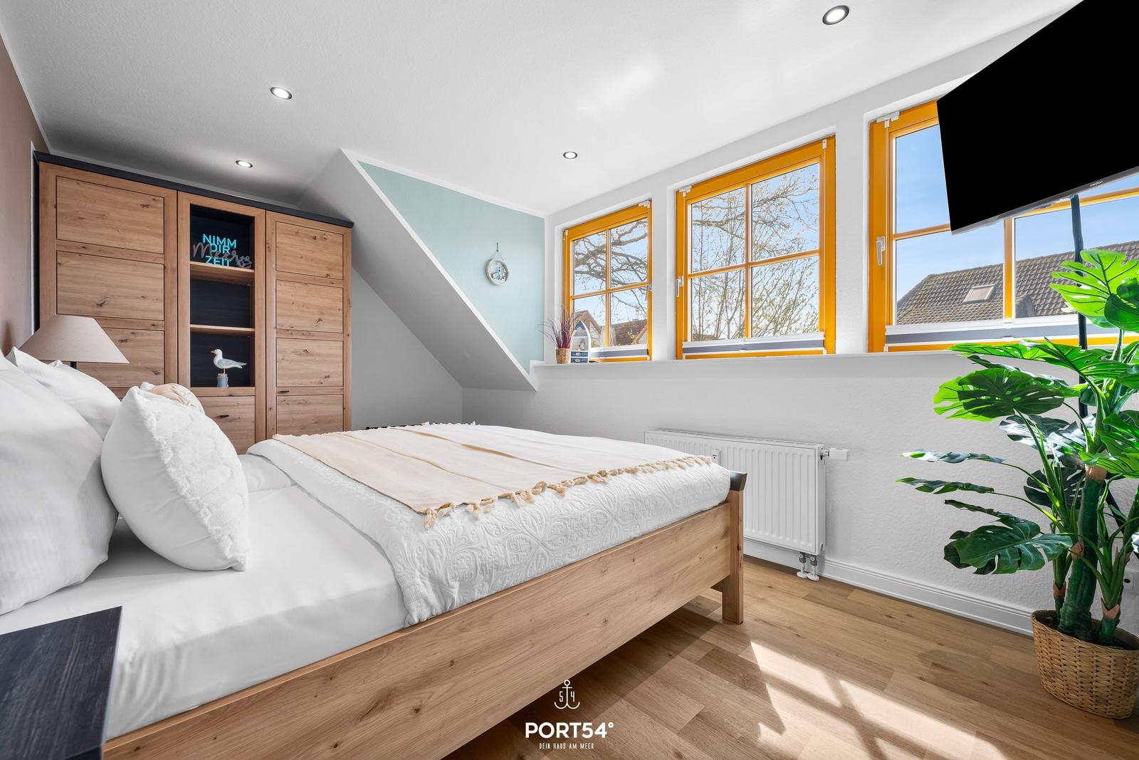 Schlafzimmer mit Holzbett, Kleiderschrank und Fenstern mit orangefarbenen Rahmen.