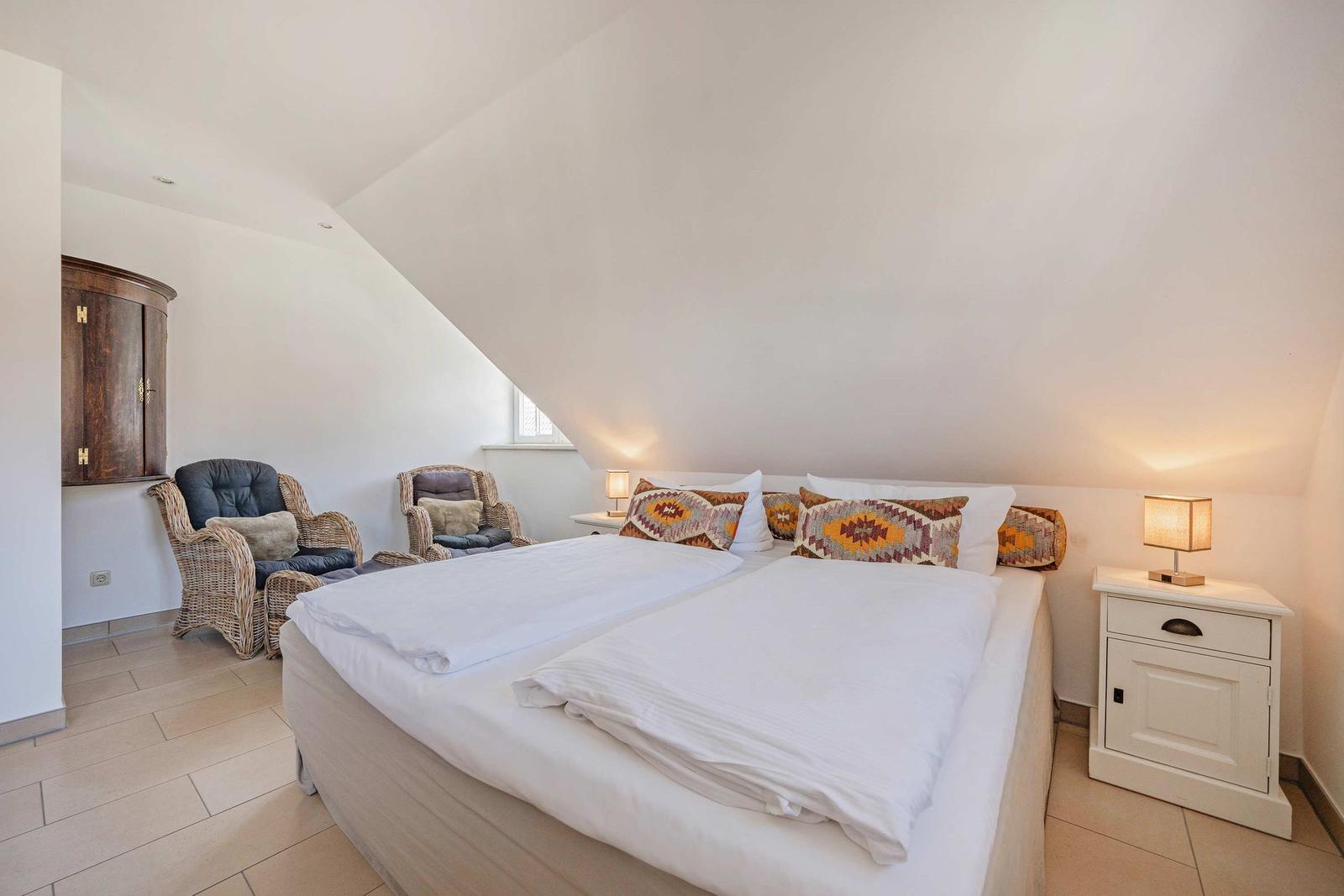 Schlafzimmer mit Doppelbett, zwei Nachttischen und zwei Sesseln unter schrägem Dach.