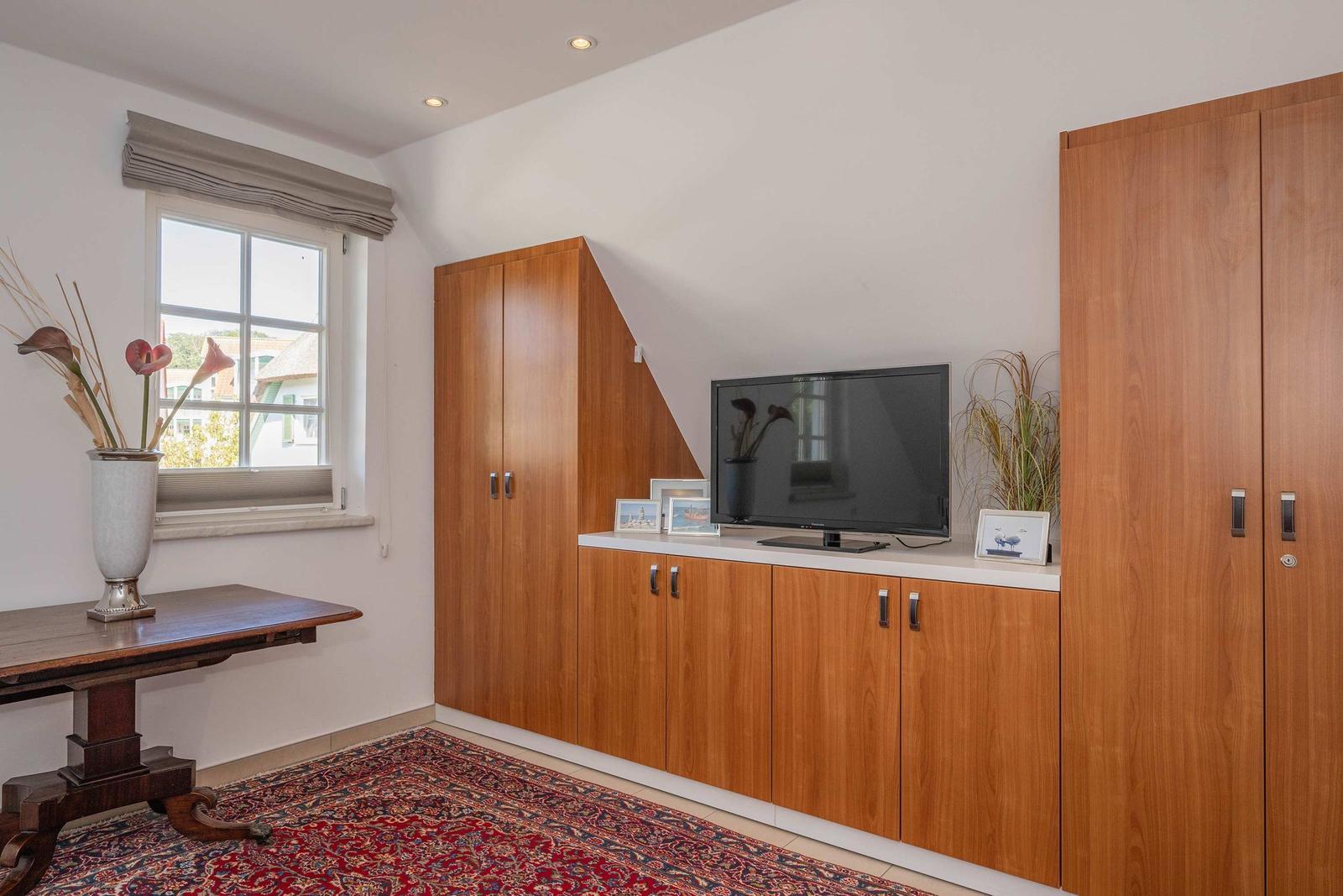 Raum mit Holzschrank, Fernseher und Fenster. Teppich und Tisch sind sichtbar.
