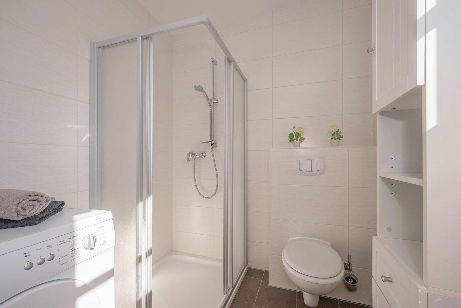Bad mit Dusche, Toilette und Waschmaschine. Weiße Fliesen und Schrank.