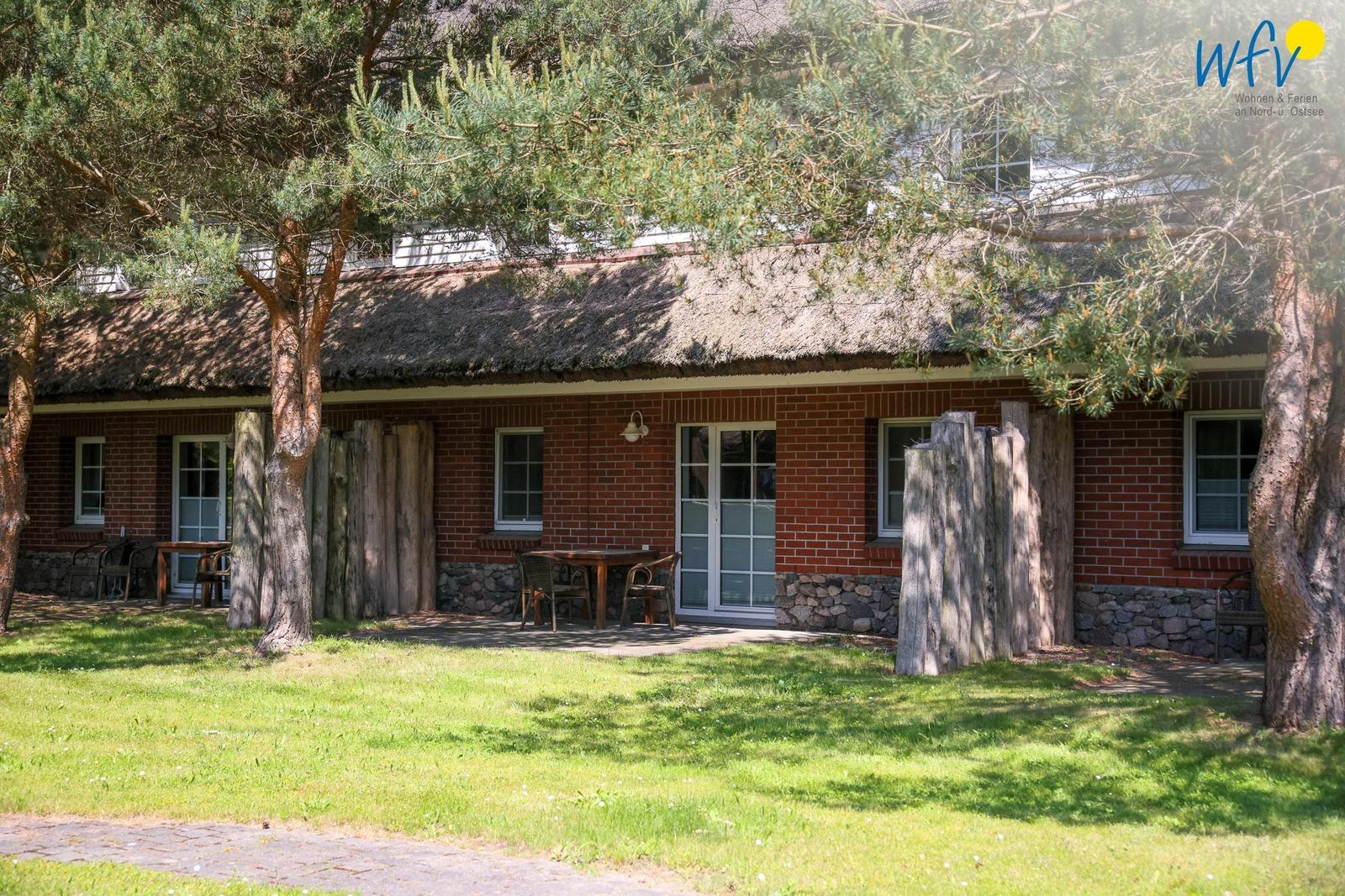 Rotes Backsteinhaus mit Strohdach, Terrasse und Bäumen. Holztische und Stühle stehen draußen.