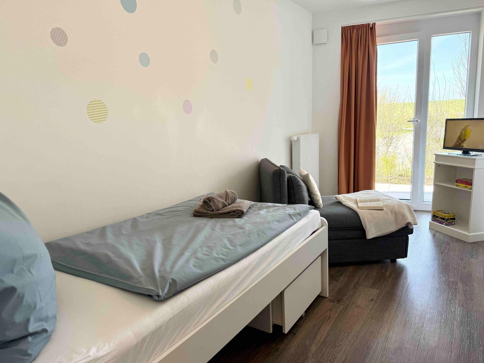 Schlafzimmer mit Bett, Sofa und TV. Durchgang zur Terrasse mit Blick auf Grünflächen.
