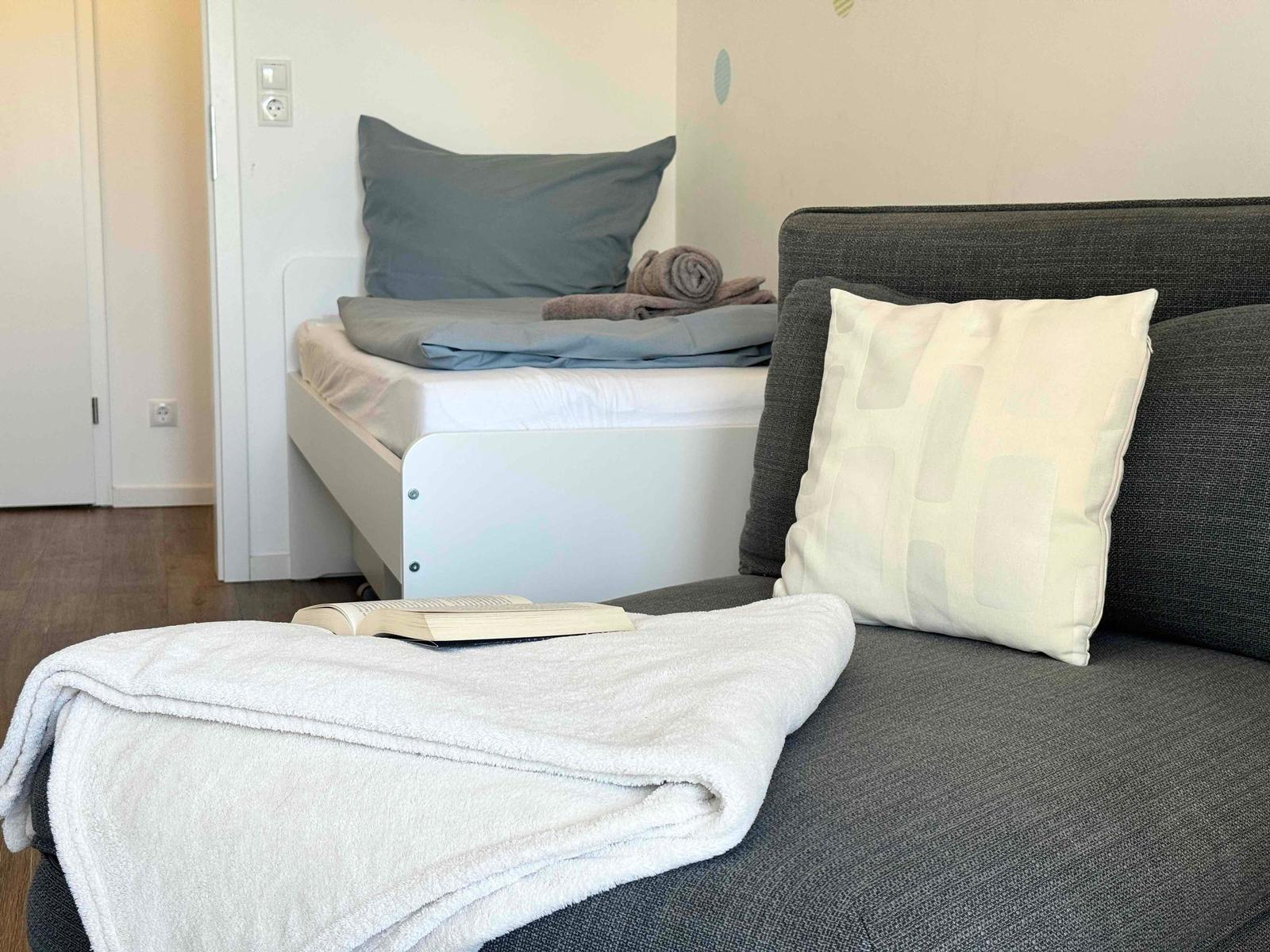 Ein grauer Sofa mit Kissen und Decke. Im Hintergrund ein Bett mit Bettwäsche.