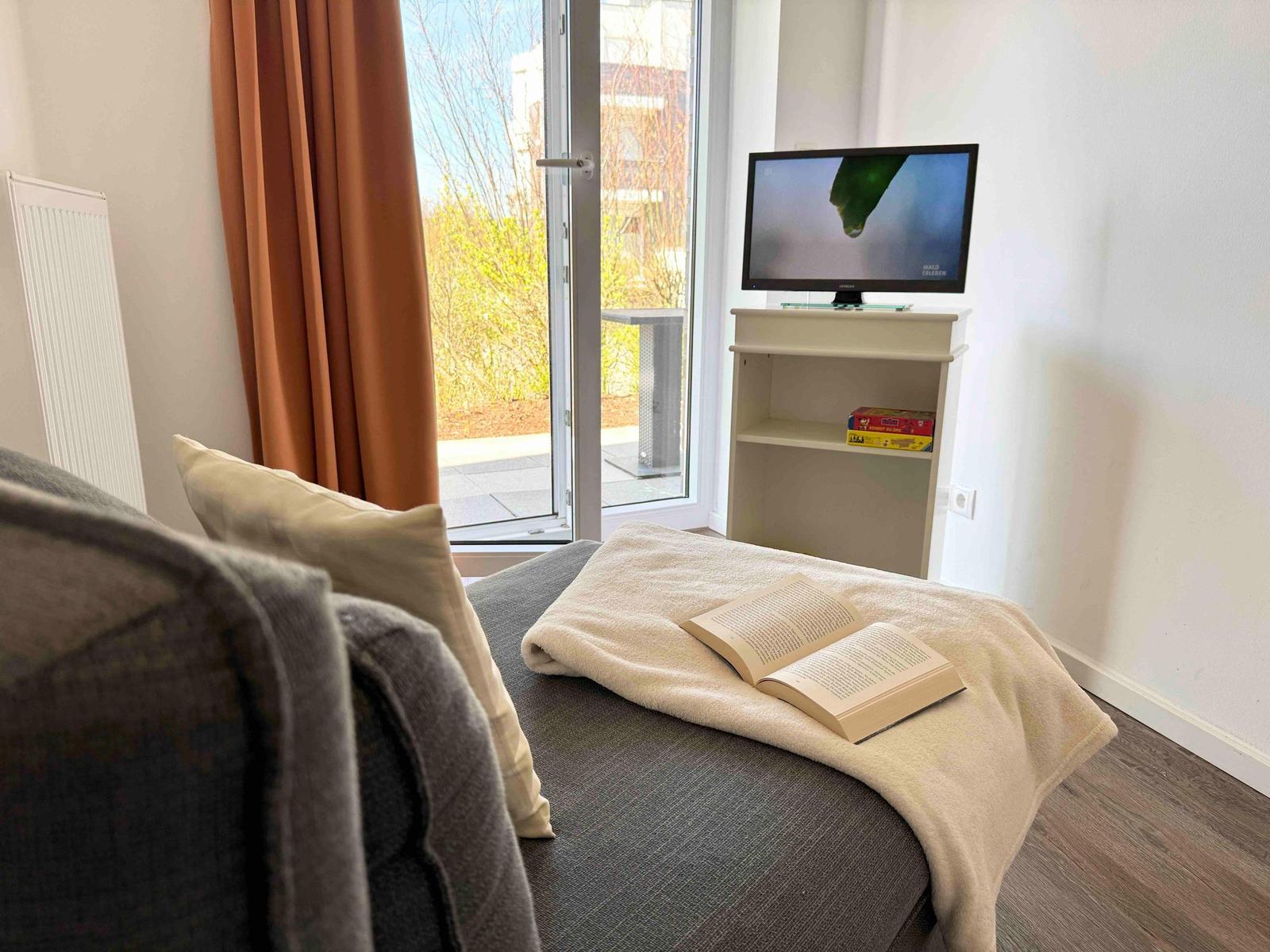Ein Sofa mit Kissen, Decke und offenem Buch. Fernseher und Balkontür.