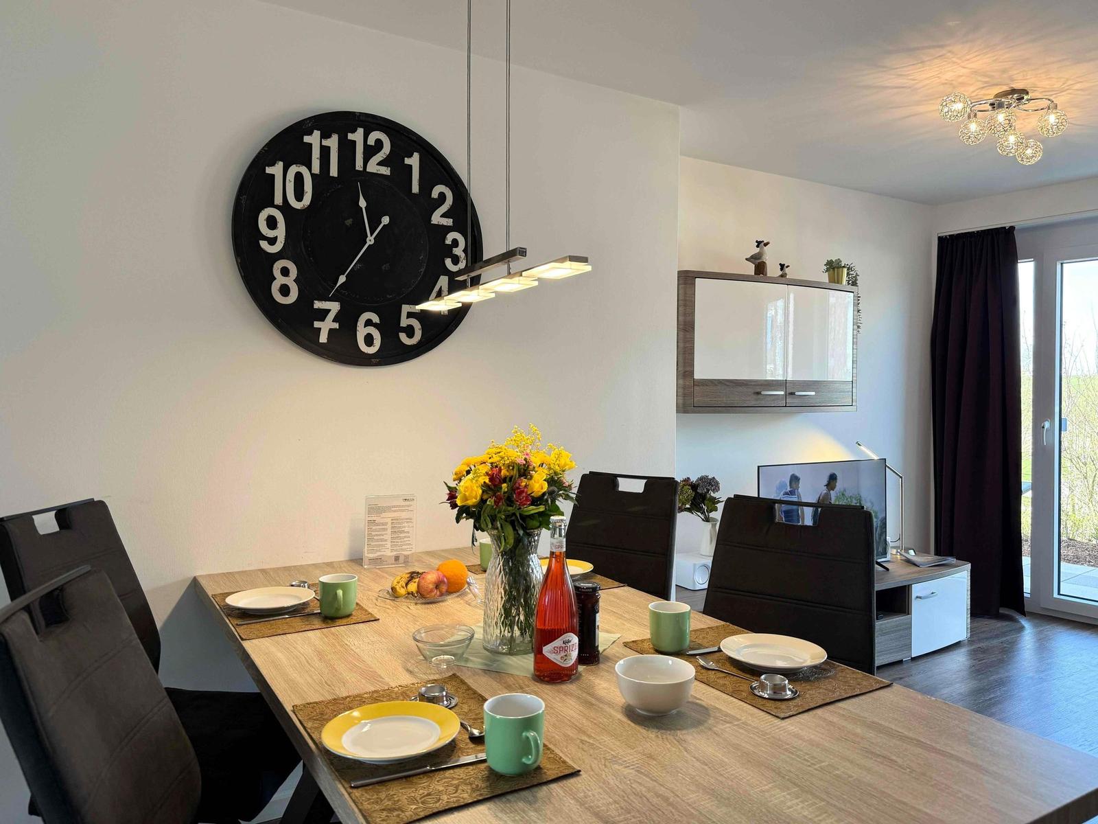 Esstisch mit Stühlen, Wanduhr und Fenster zur Terrasse.
