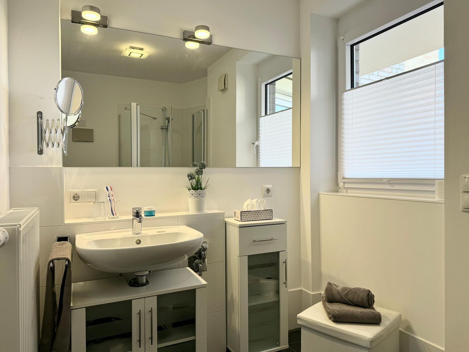 Badezimmer mit Waschbecken, Spiegel, Dusche und Fenster mit Jalousie.